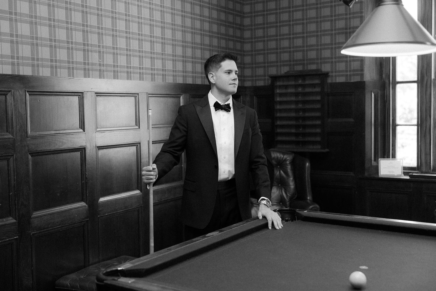 Man in tuxedo holding pool cue beside billiard table in elegant wood-paneled room, black and white photo.