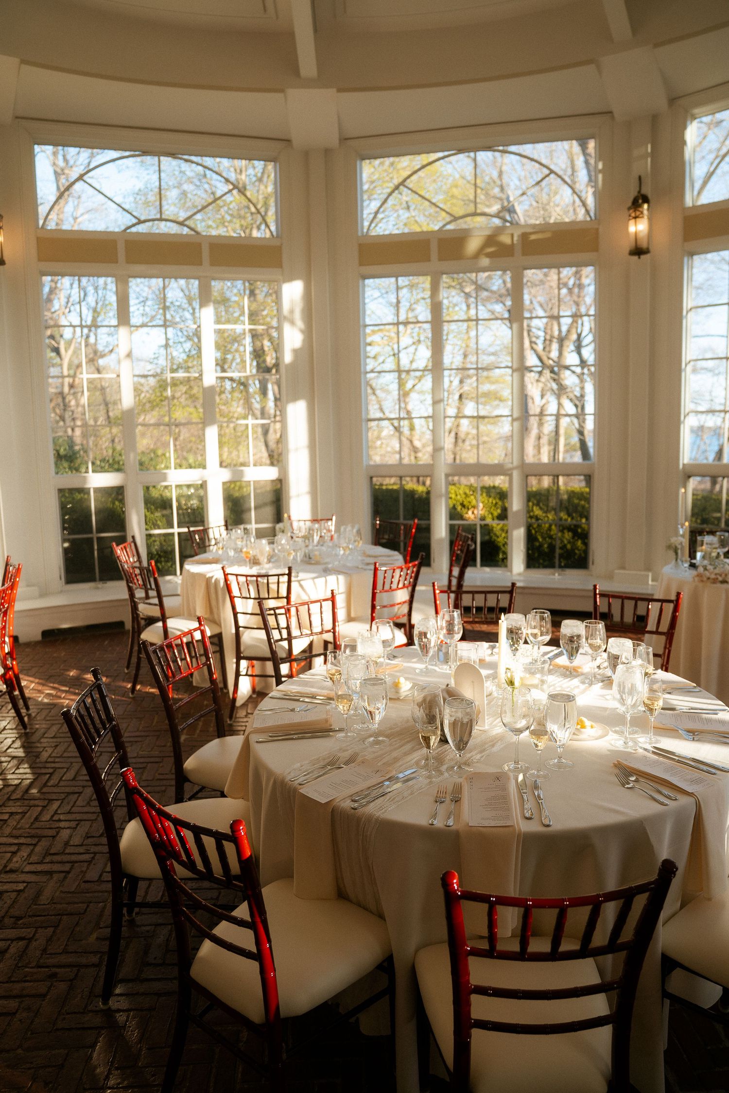 Elegant wedding reception venue with round tables set with white linens and chiavari chairs in a sunlit ballroom.