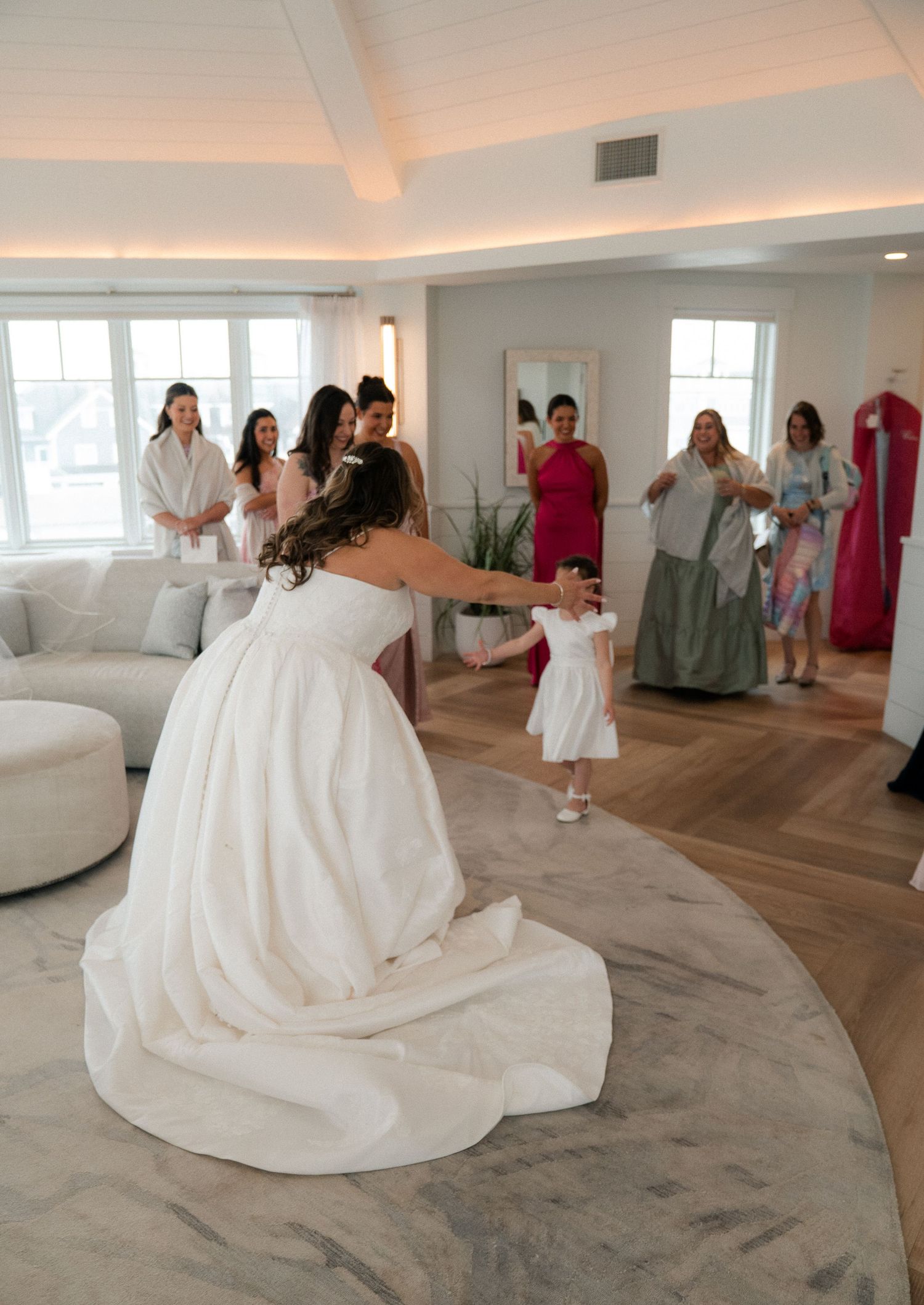 Bride in white ball gown kneeling to embrace a small flower girl while bridesmaids watch joyfully.