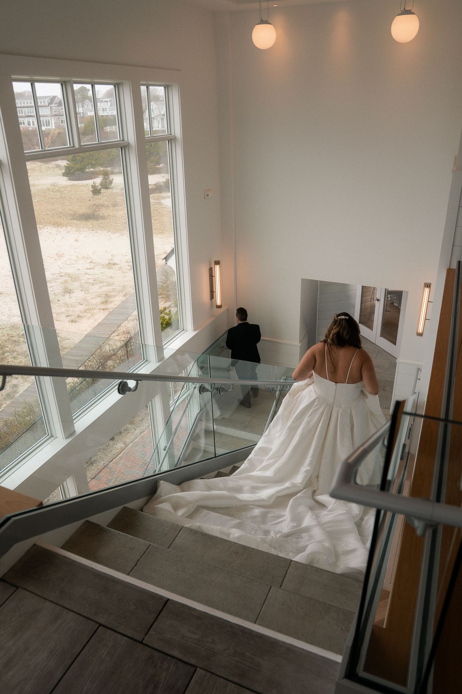 Bride in white gown ascending modern staircase with glass railings, groom ahead, large windows overlooking sandy landscape.