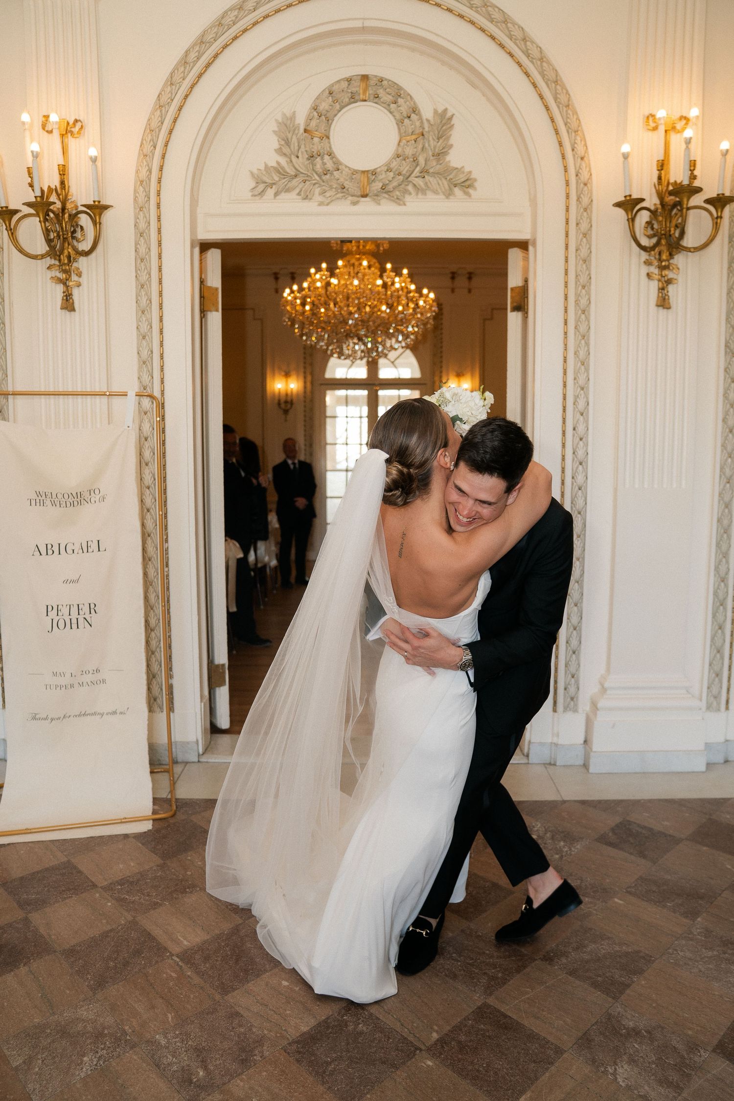 Newlyweds embrace and laugh joyfully in a grand ornate venue doorway, bride wearing a backless white gown and veil.