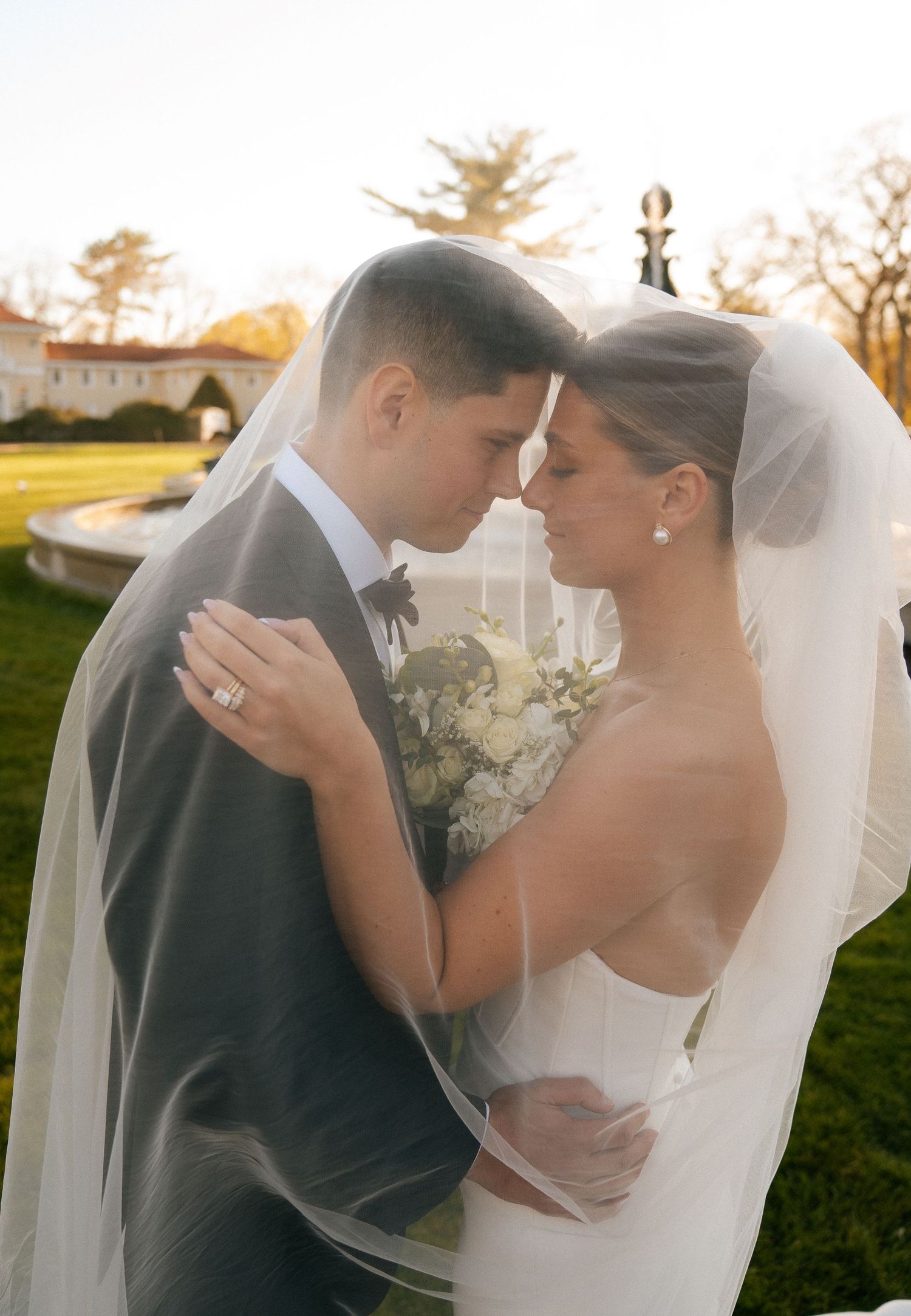 Bride and groom share an intimate moment under a flowing wedding veil outdoors at golden hour.
