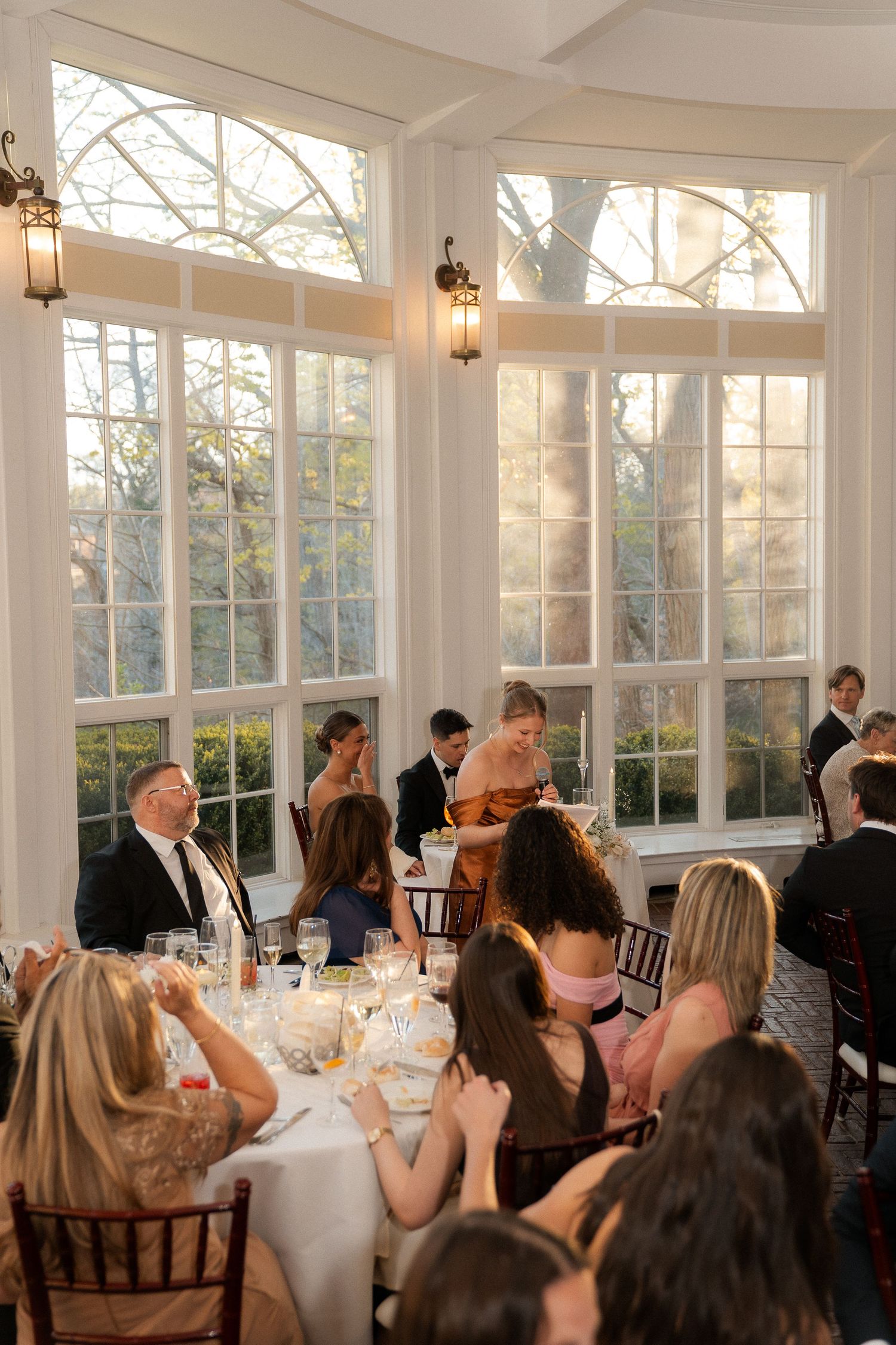 Elegant wedding reception in a sunlit ballroom with large windows, guests seated at round tables with white linens.