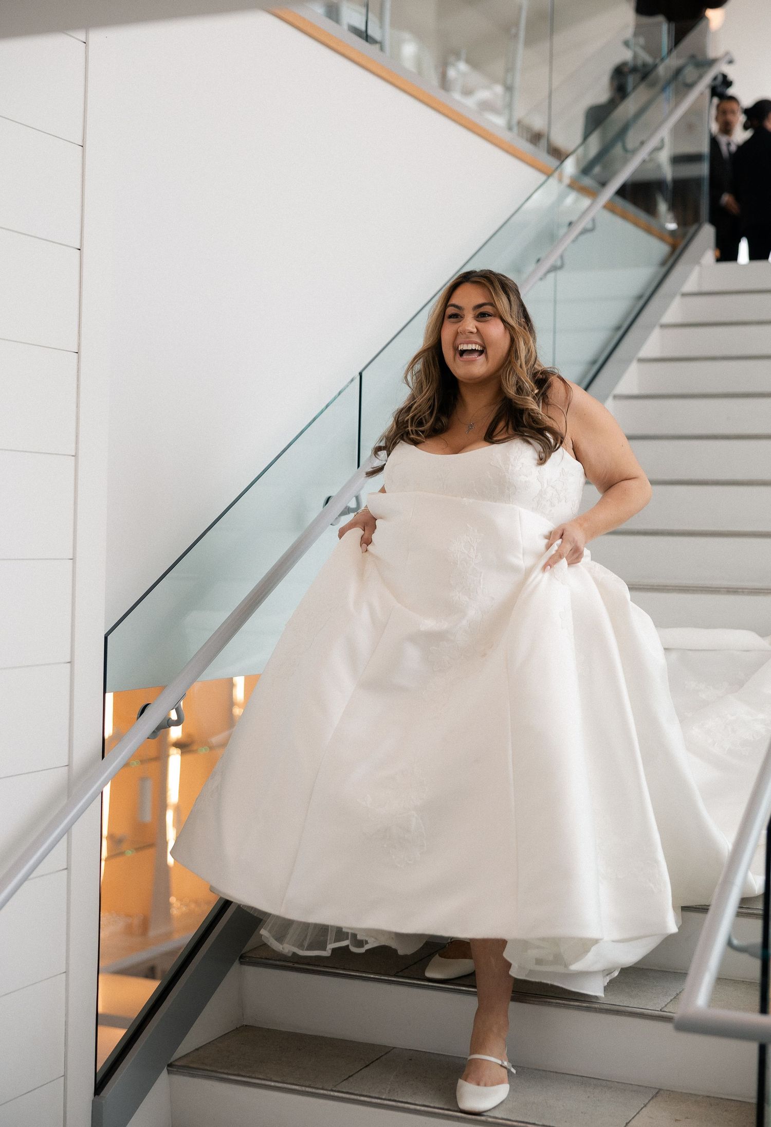 Laughing bride in white ball gown holding dress hem on modern staircase with glass railing.