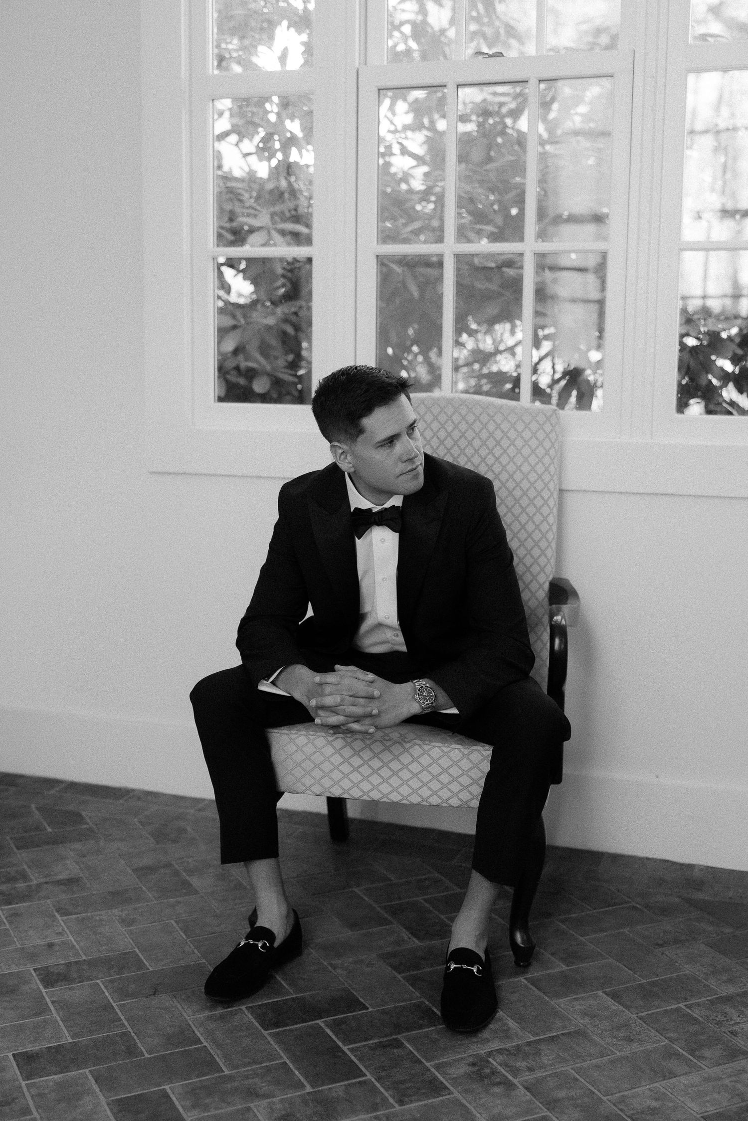 A groom in a black tuxedo and bow tie sits elegantly in a chair by a window in a black and white photo.
