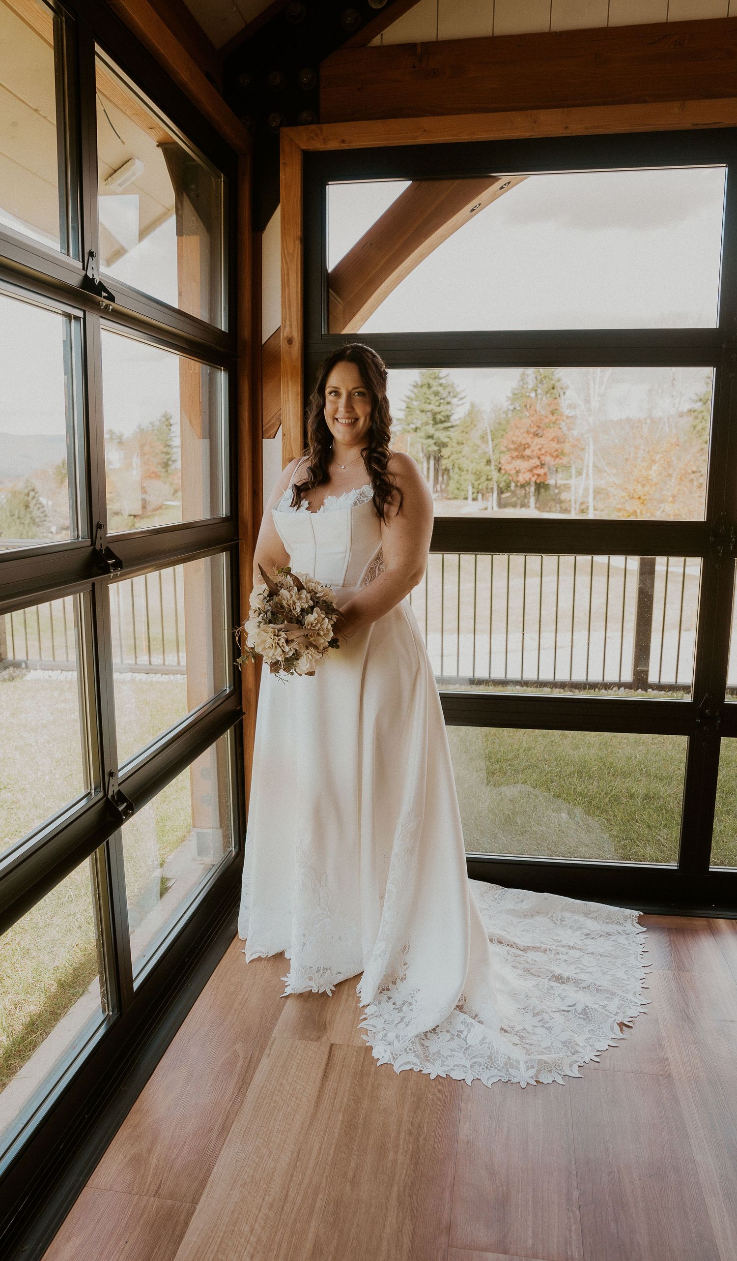 A white lace wedding dress displayed in a sunlit room with wooden floors and large windows.