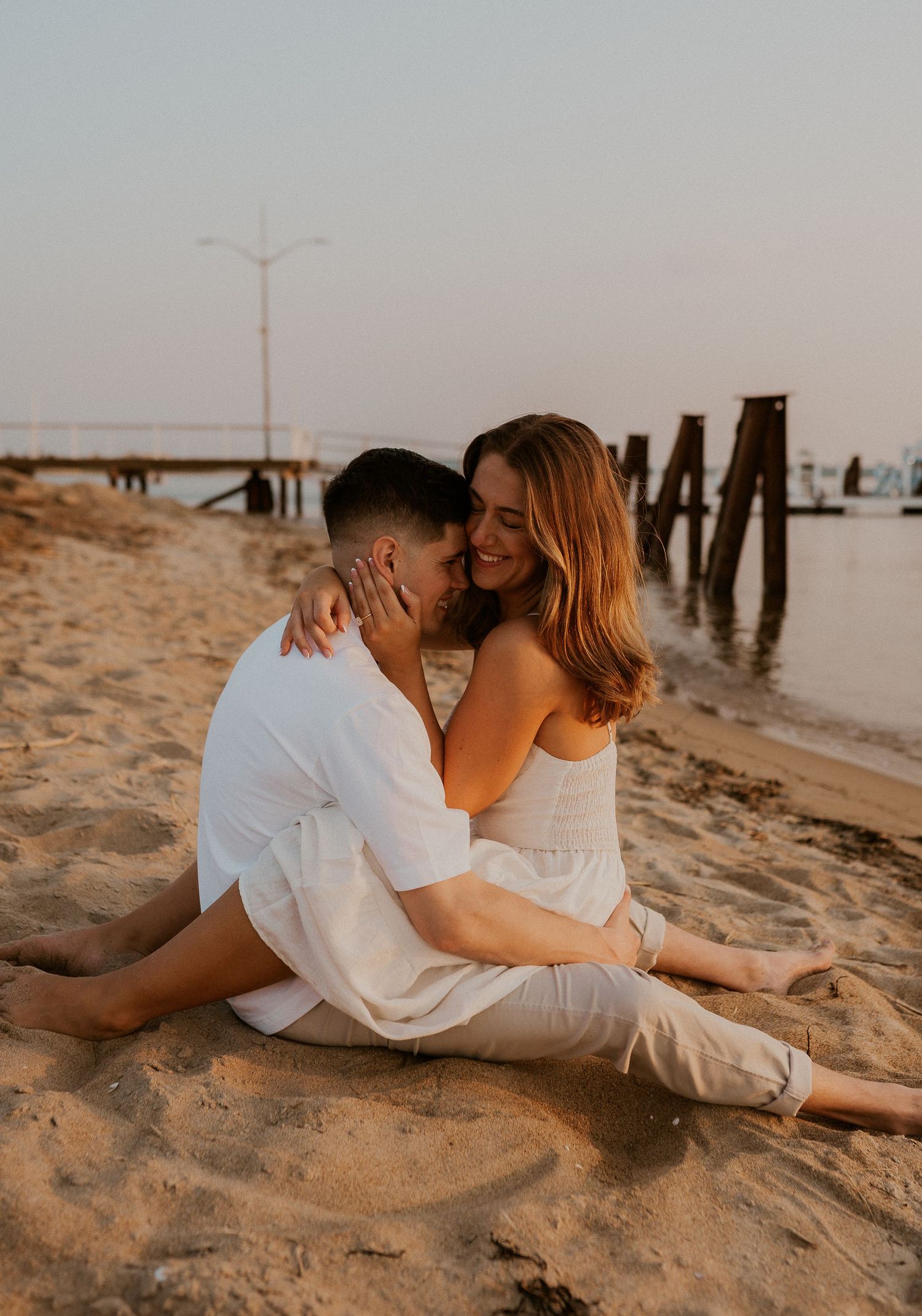 Romantic couple sitting on beach sand near wooden pier posts during golden hour.