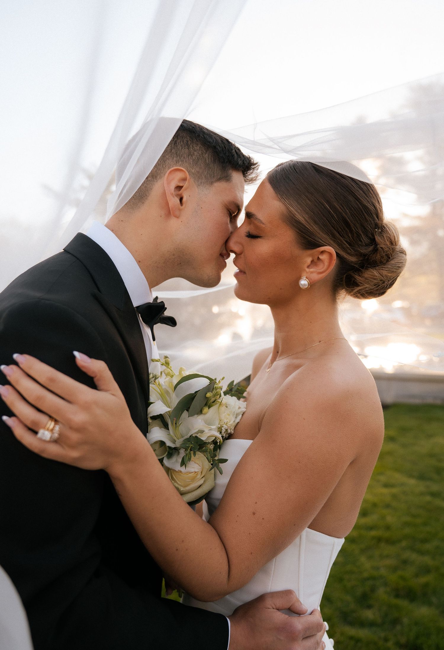 Bride and groom sharing a romantic kiss under a veil, bride holding white floral bouquet at golden hour.