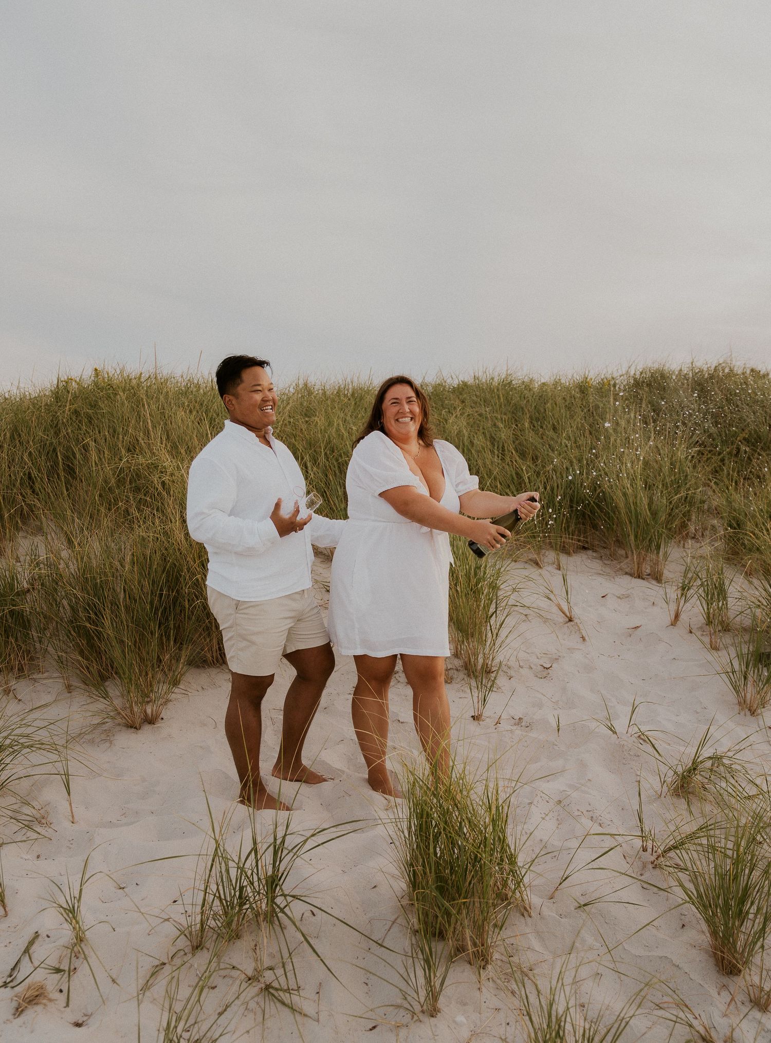 Couple in white outfits celebrating on sandy beach dunes, popping champagne bottle with tall grass in background.