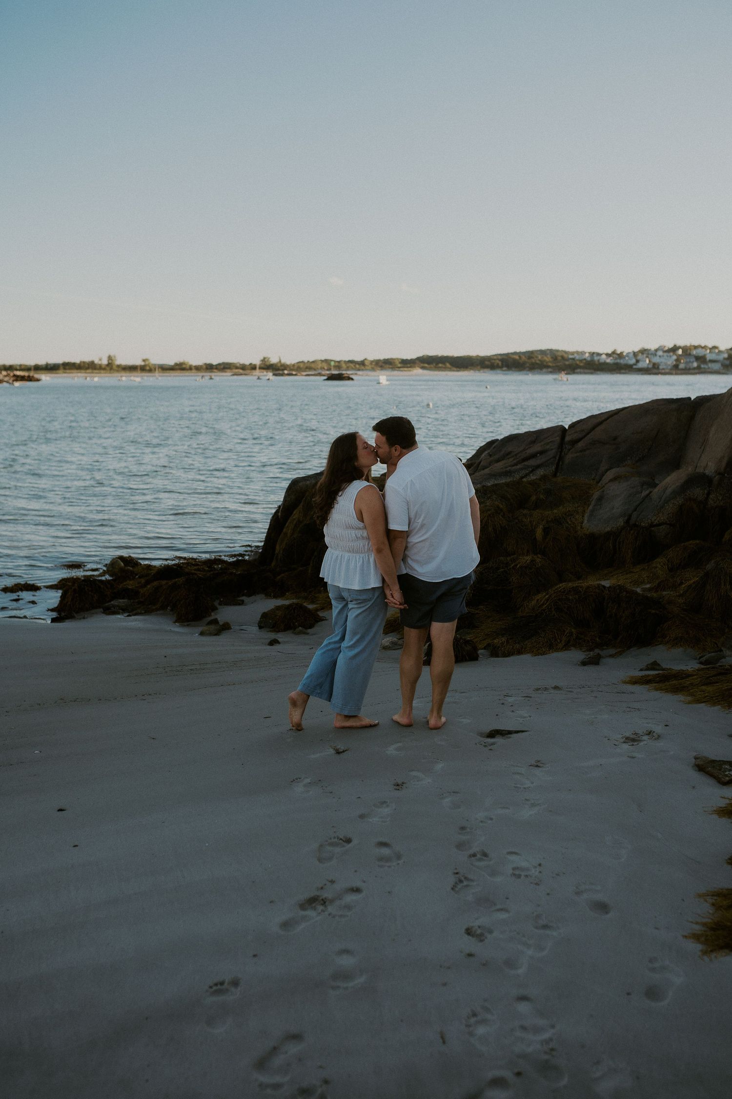 Two people exploring a rocky beach at dusk with boats visible in the harbor behind them.