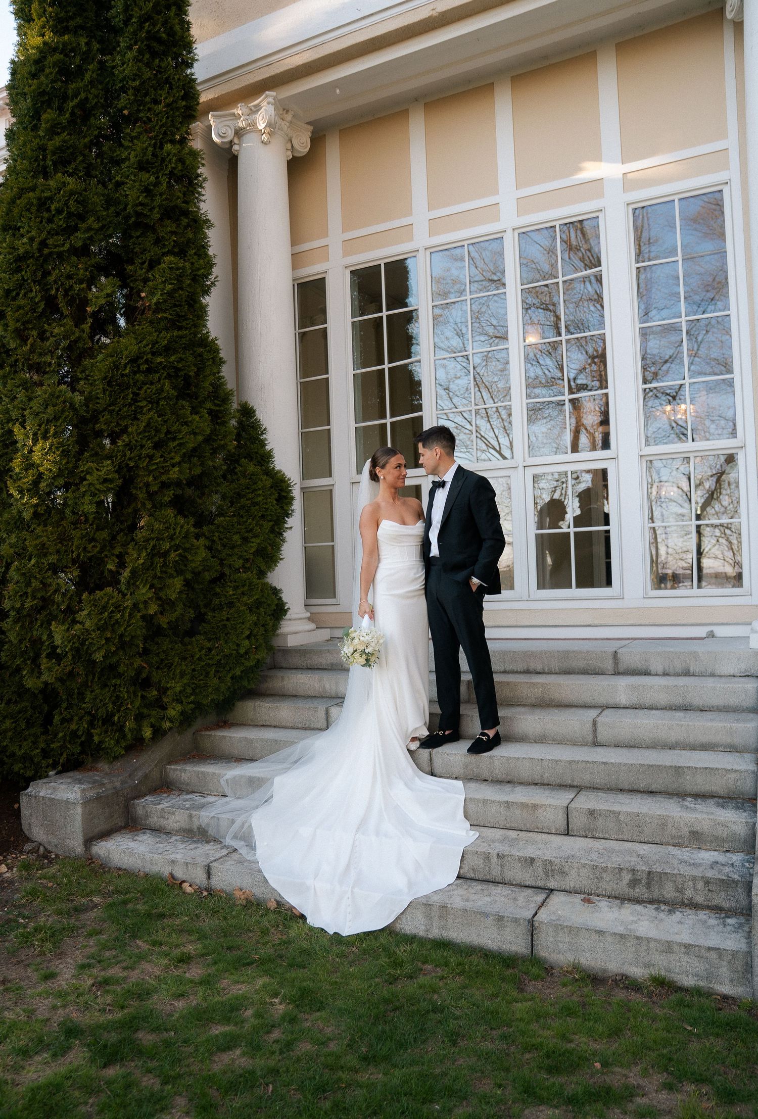 Bride in strapless gown with long train poses with groom on stone steps of elegant venue.