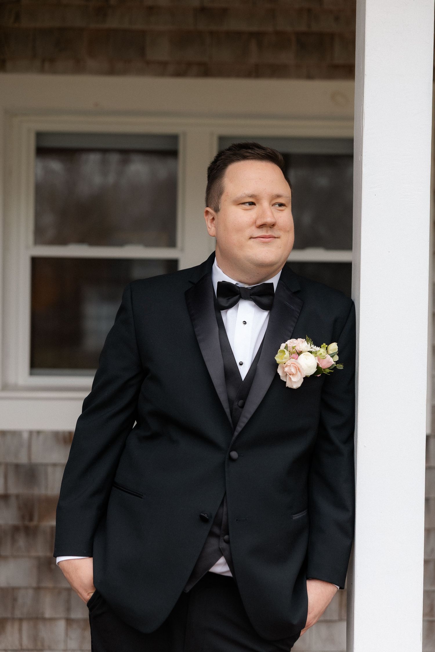 Dapper groom in black tuxedo with floral boutonniere leaning against a white column outdoors.