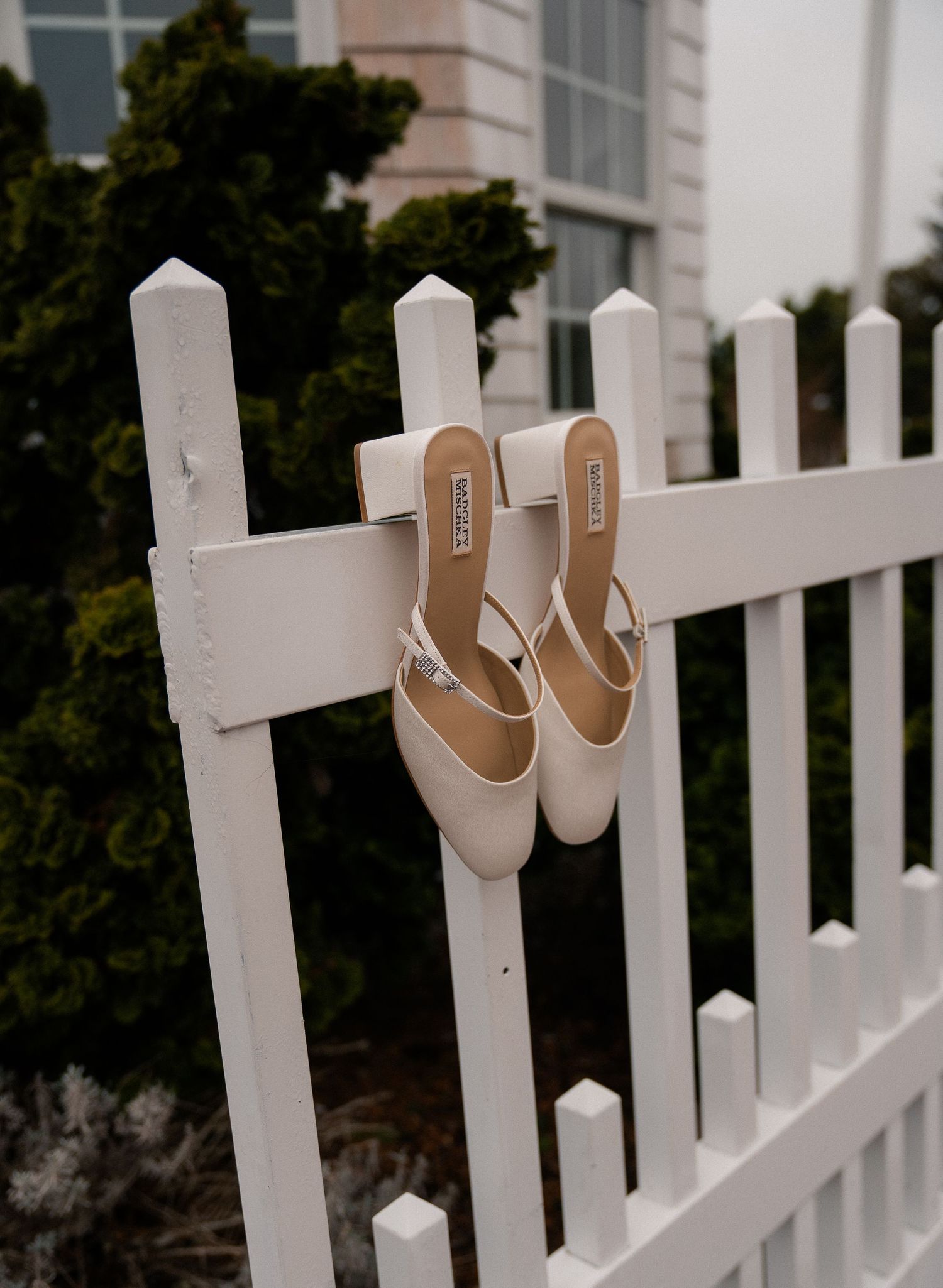 White bridal heels hanging on a white picket fence with green shrubs in the background.