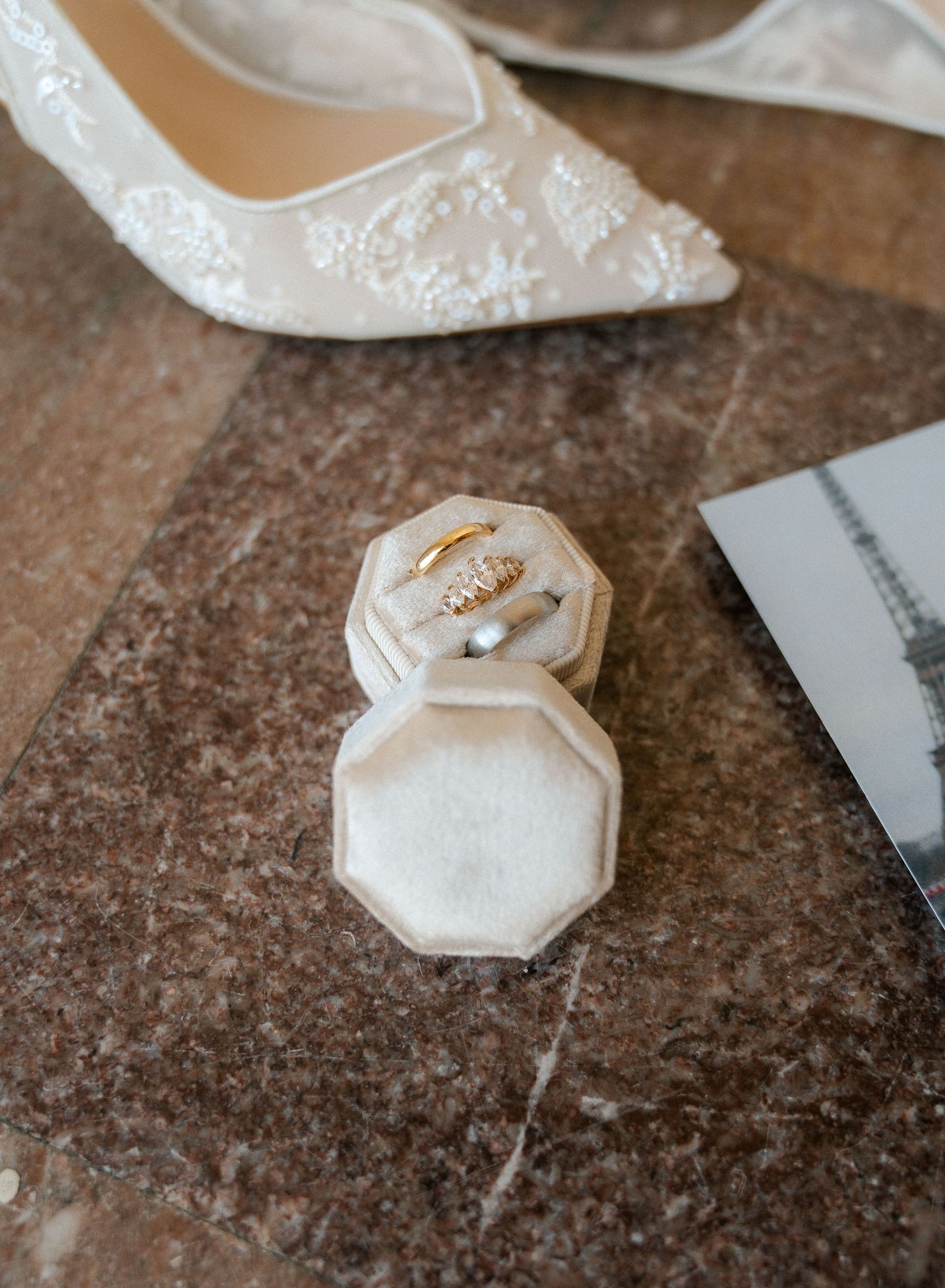 Elegant wedding rings displayed in a velvet octagonal ring box beside lace bridal heels on a marble surface.