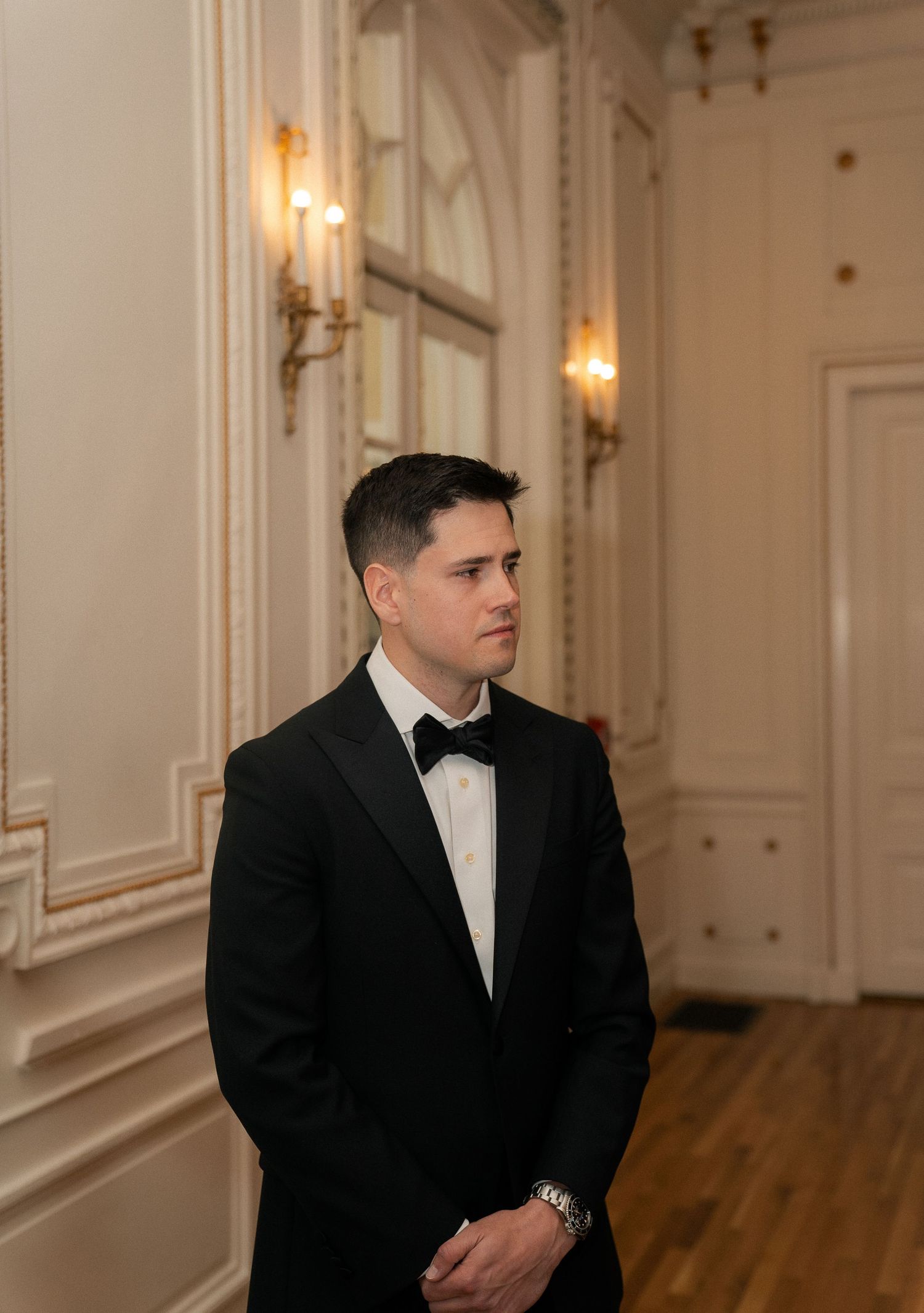 A man in a black tuxedo with bow tie stands in an elegant white room with ornate moldings and gold wall sconces.