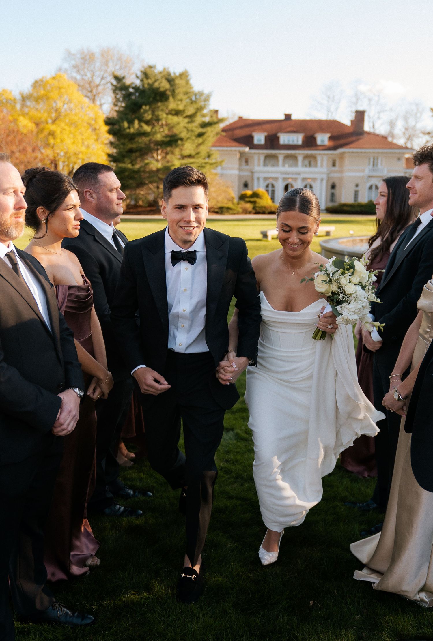 Bride and groom walk hand-in-hand through wedding party outdoors at elegant estate, bride in strapless gown holding bouquet.