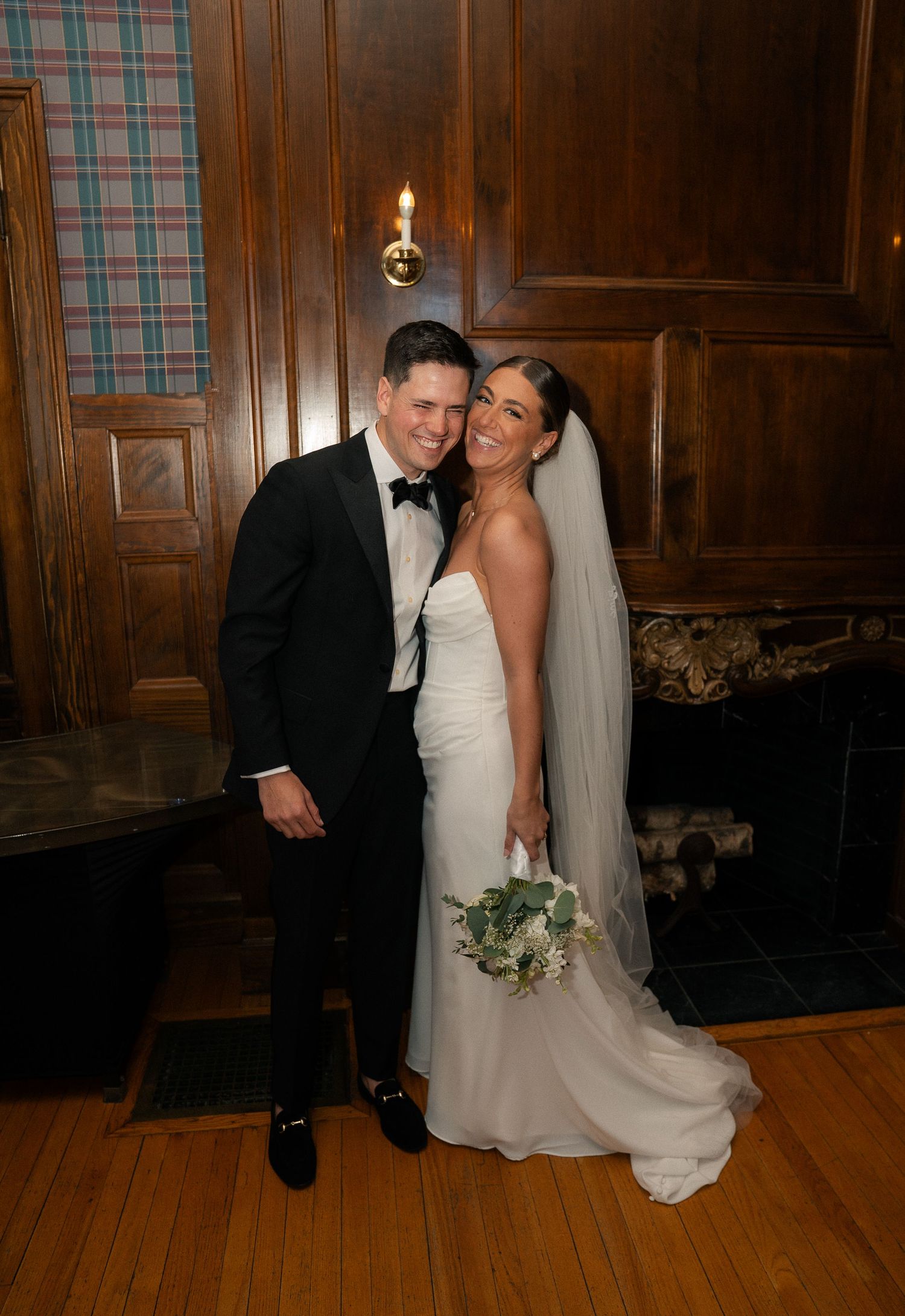 Bride and groom posing together in elegant wood-paneled room, bride in strapless white gown with veil holding bouquet.