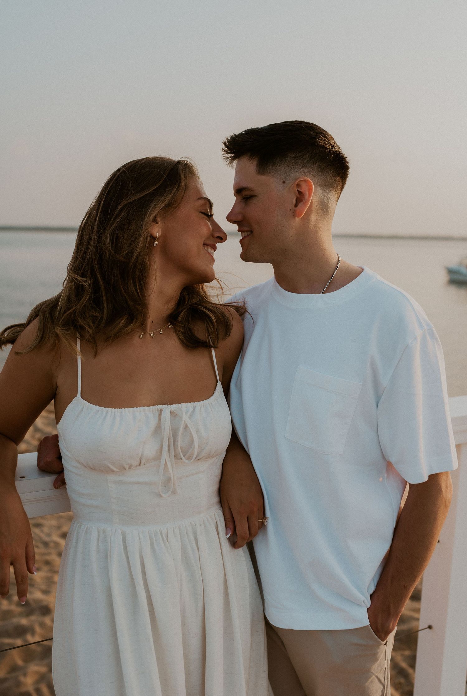 A romantic couple in white outfits share an intimate moment at sunset by the waterfront.
