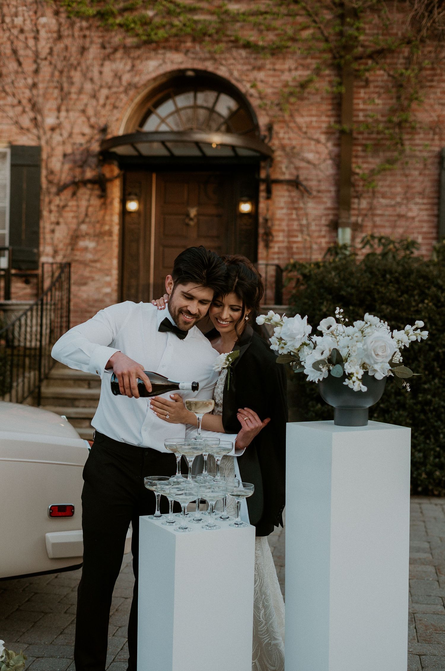 Wedding reception setup with white pedestals and floral arrangements against brick wall.