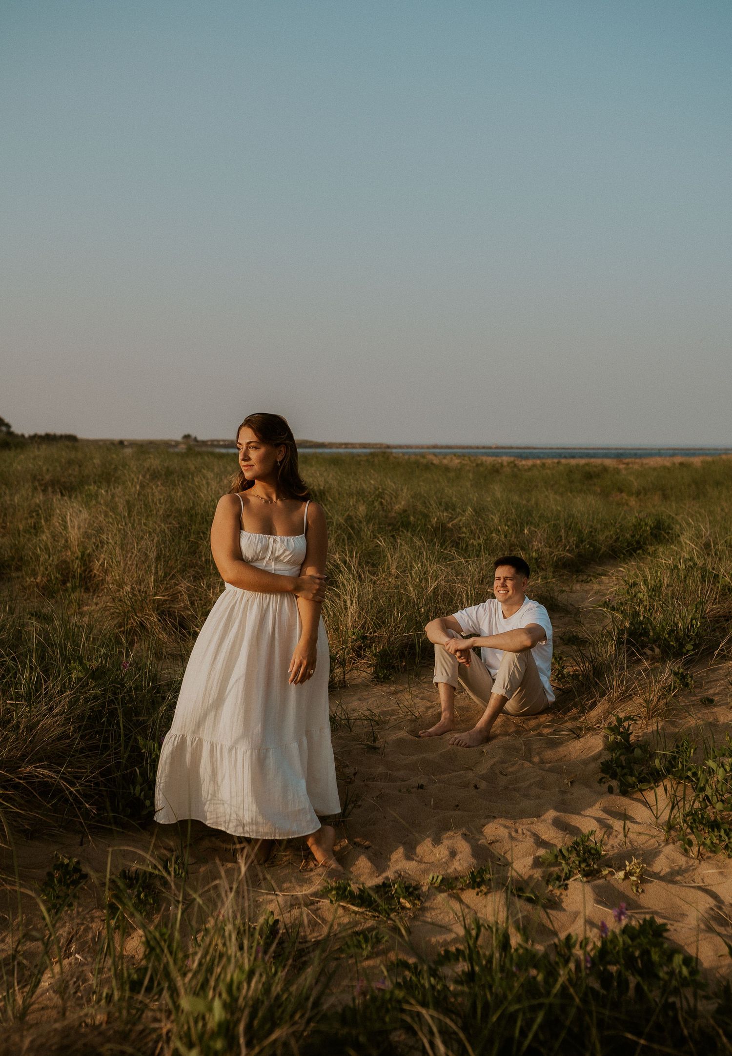 Two people in white dresses sit and embrace on sandy dunes at sunset with grassy fields and ocean in background.