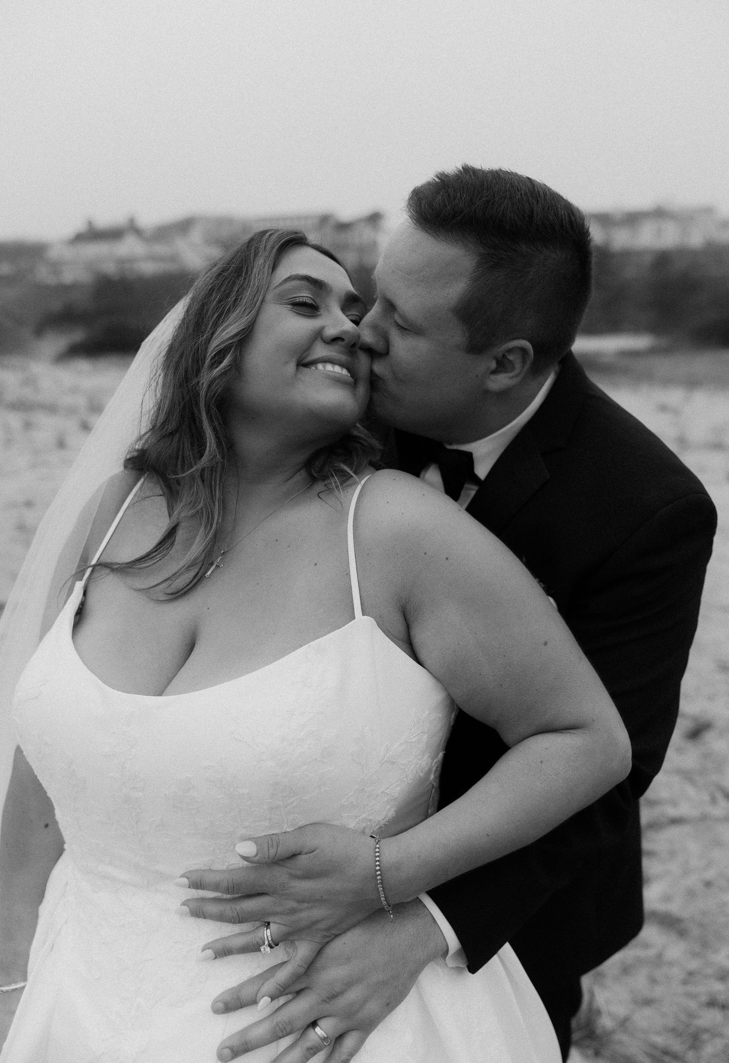 Black and white photo of a groom kissing his bride on the cheek at a beach wedding, bride smiling in white gown and veil.