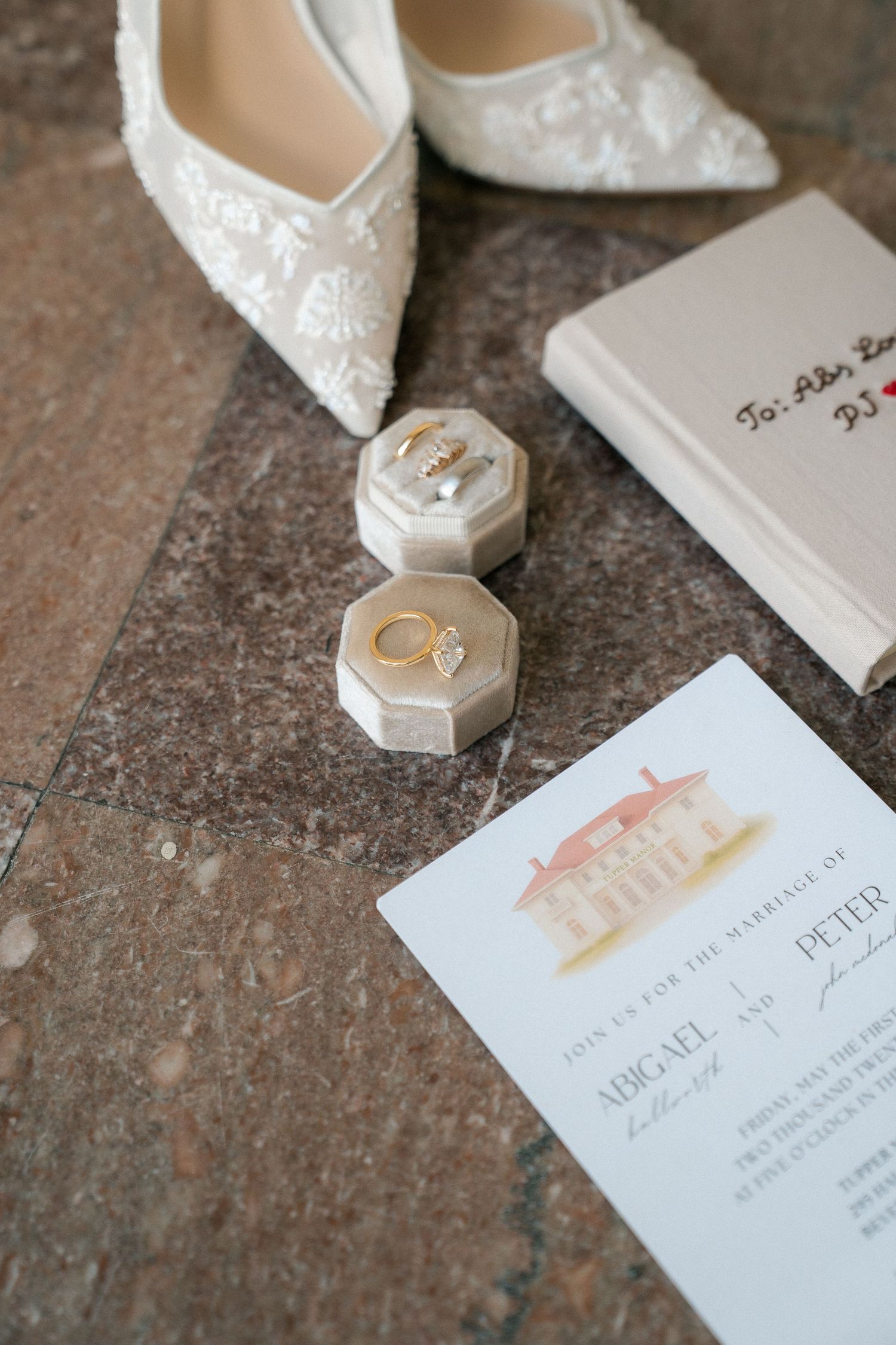 Elegant wedding flat lay featuring lace bridal heels, gold rings in velvet box, pearl earrings, and watercolor venue invitation on marble.