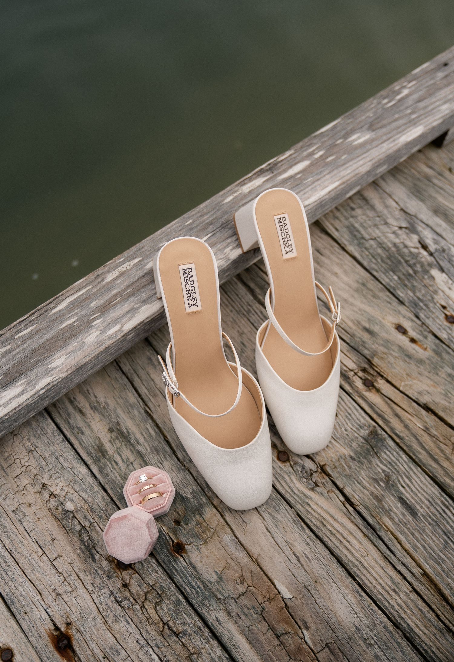 White block-heel bridal mules by Badgley Mischka rest on a weathered dock beside a pink ring dish.