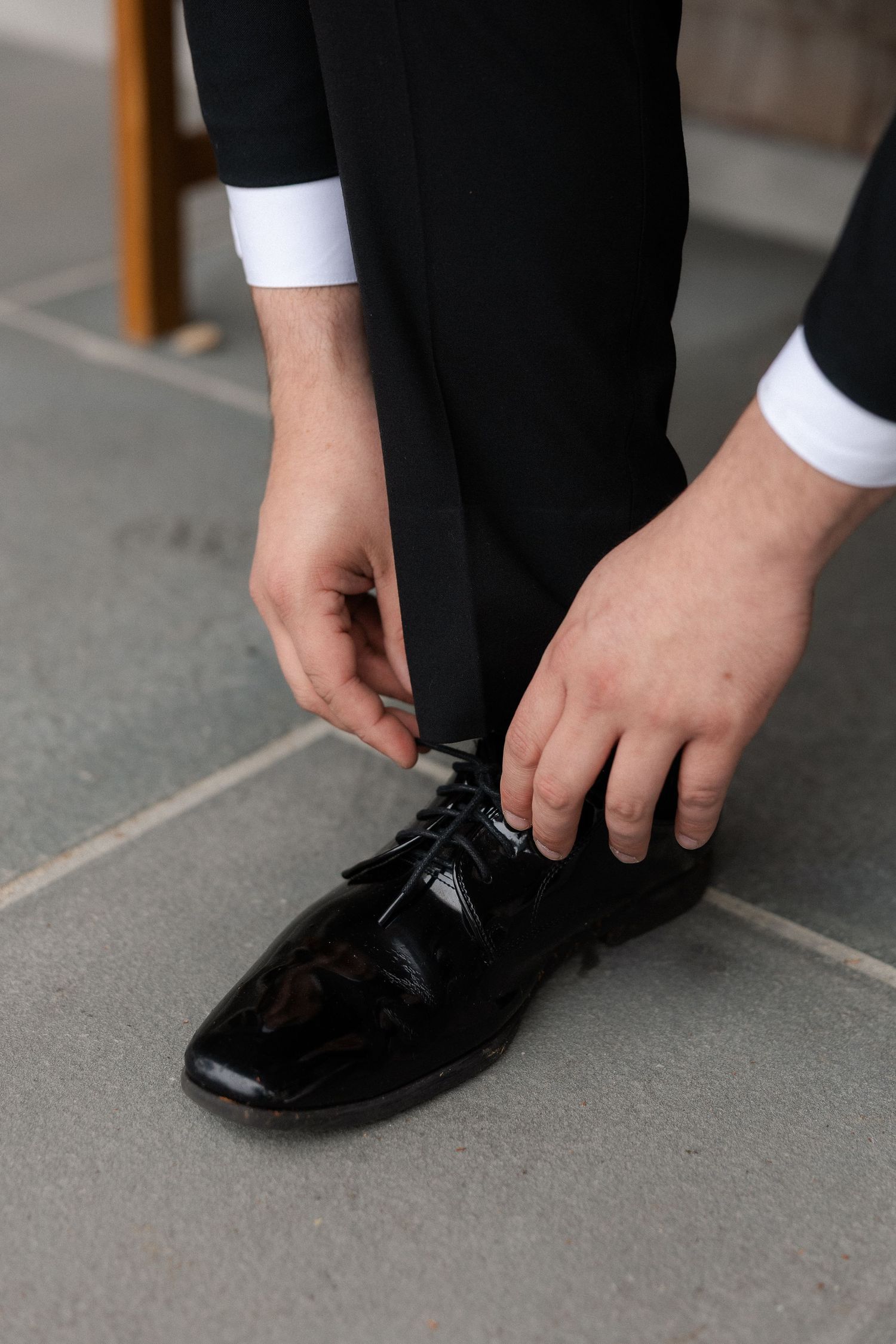 Groom in black tuxedo tying shiny patent leather dress shoes on a stone patio before the wedding.