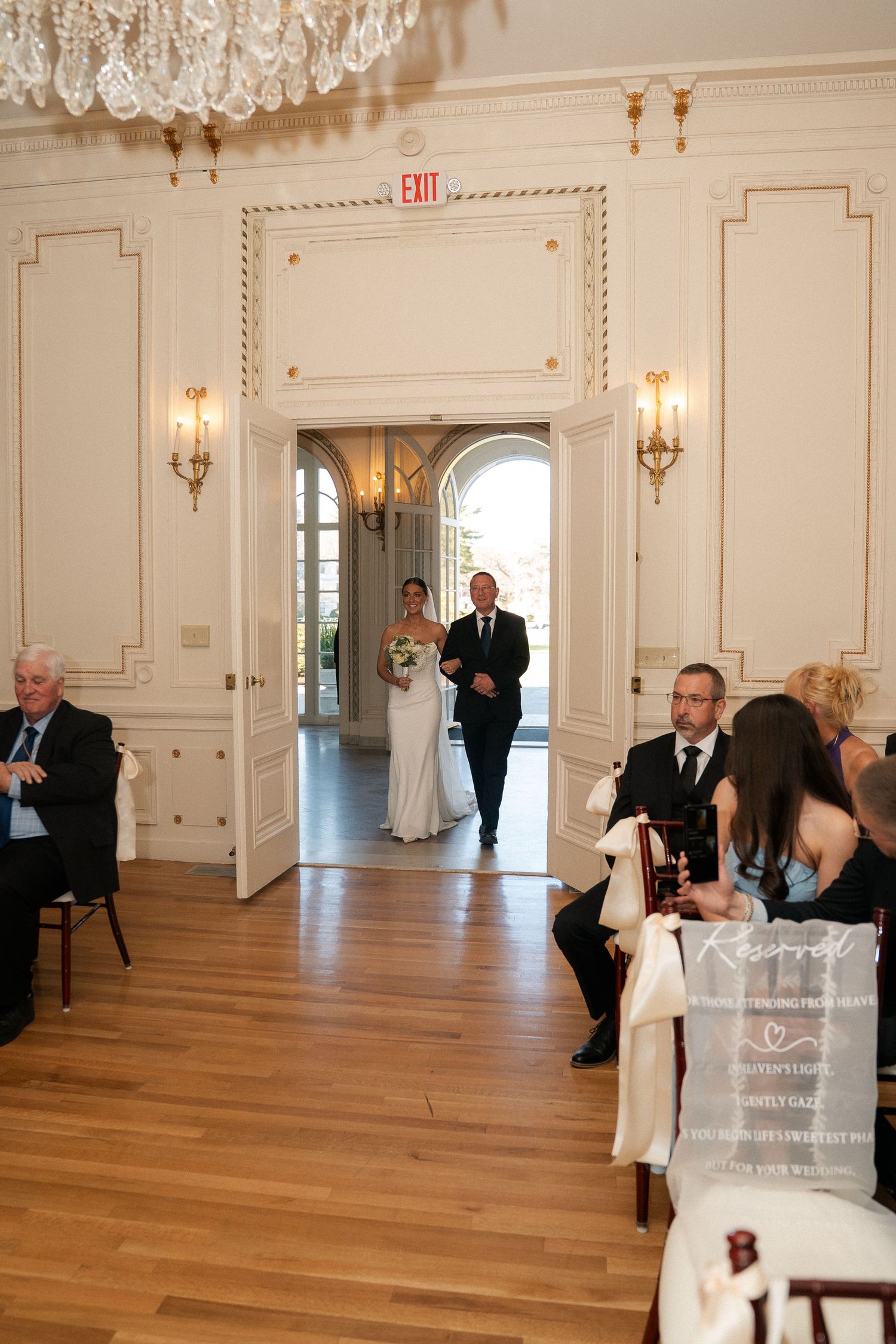 Bride walks down the aisle with escort in elegant white ballroom during wedding ceremony.