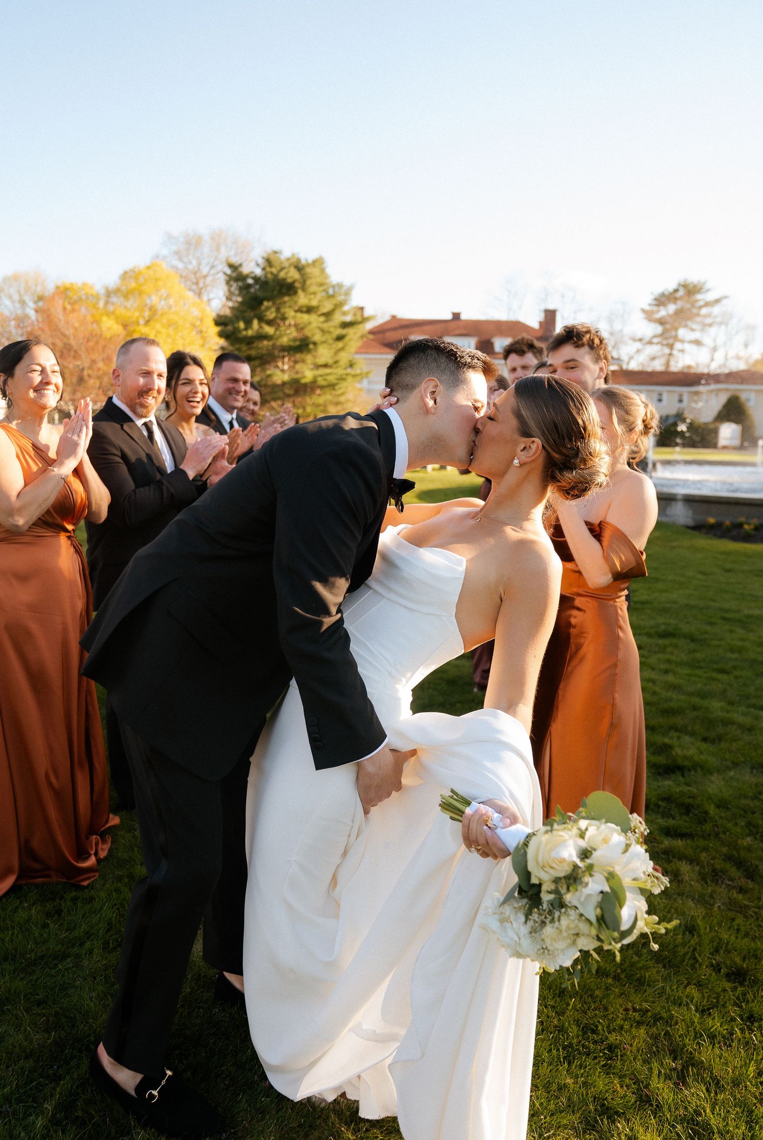 Bride and groom share a kiss surrounded by wedding party in rust bridesmaid dresses on a sunny outdoor lawn.