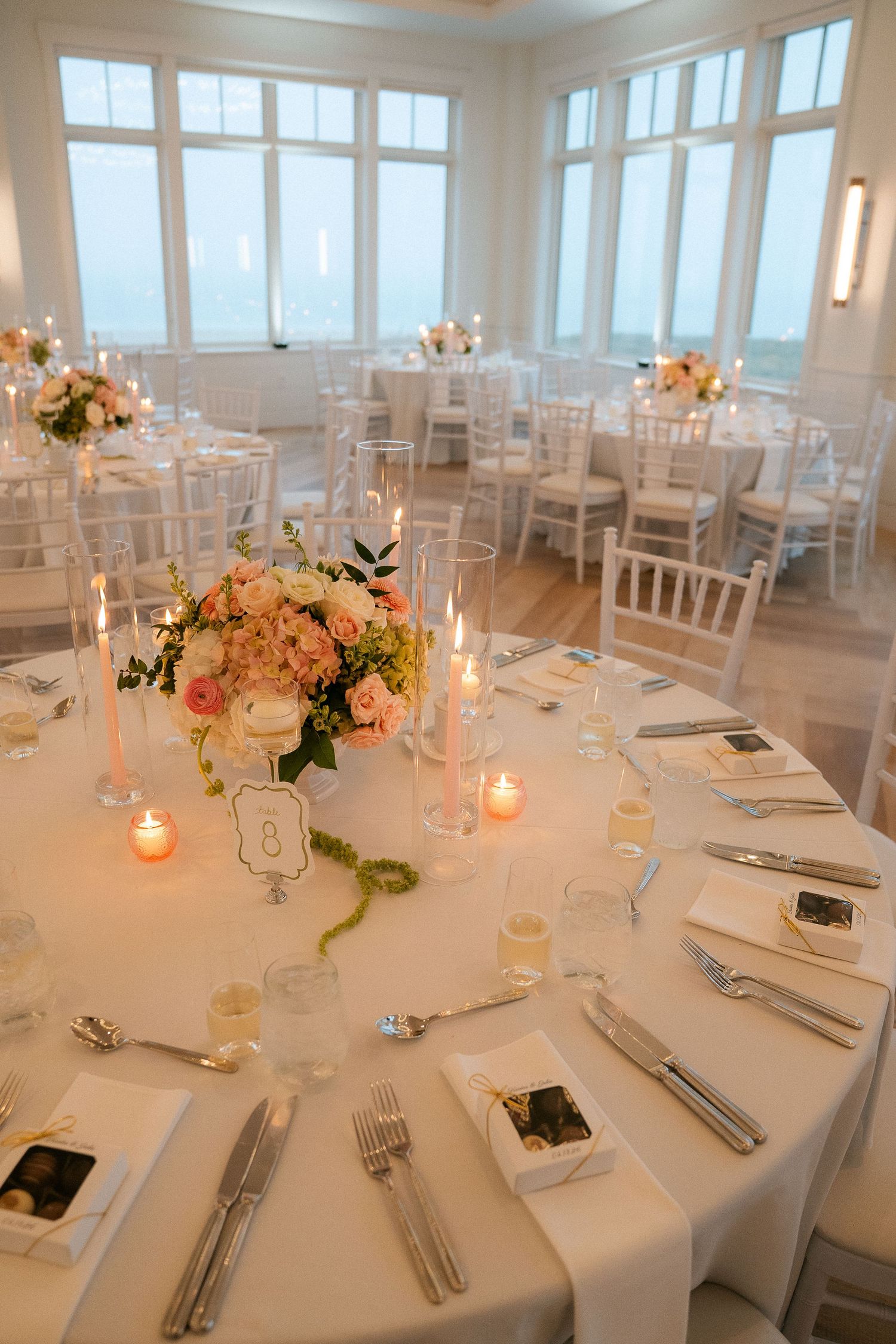 Elegant wedding reception table set with pink floral centerpiece, taper candles, and white linens by large windows.