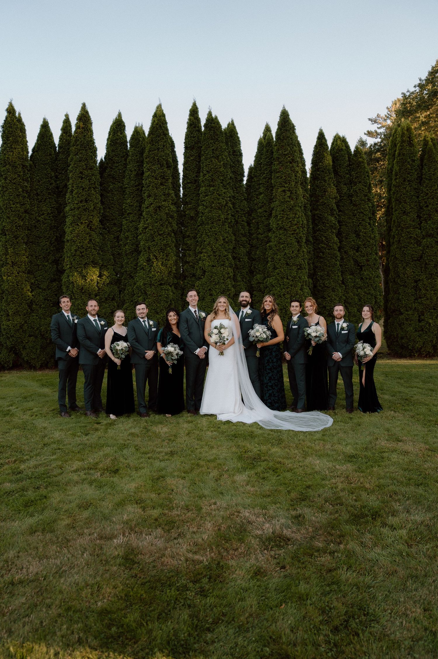 Wedding party in black attire poses with couple against a backdrop of tall evergreen trees.
