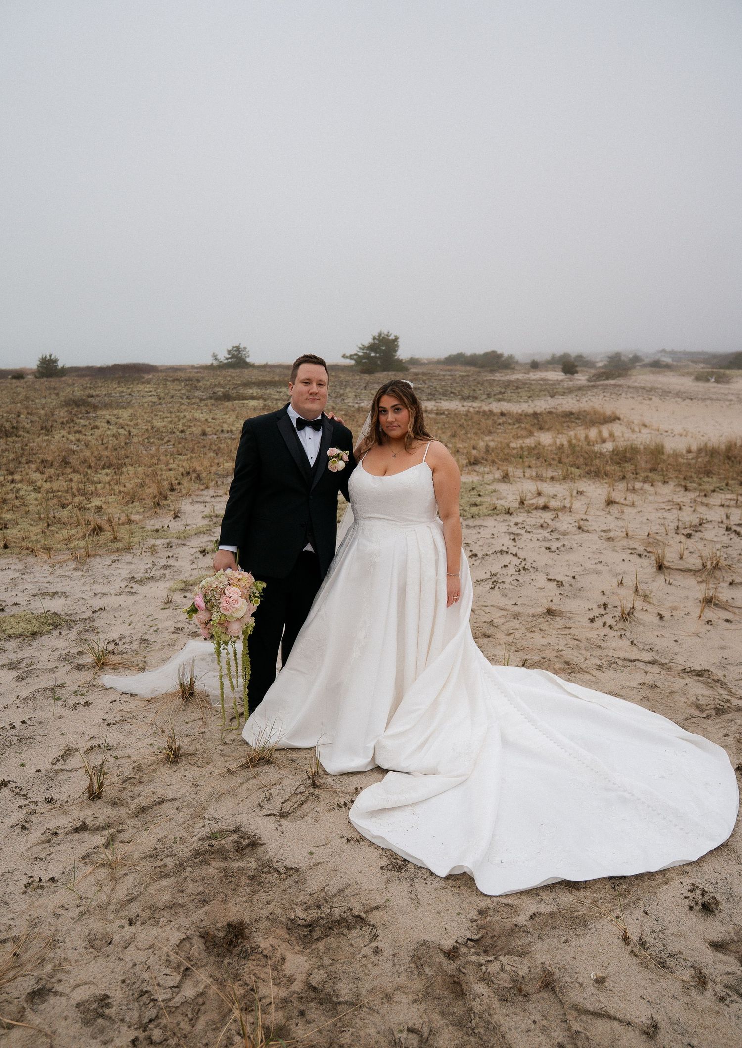 Bride and groom posing on sandy dunes, bride in white gown with train, groom in black tuxedo with bow tie.