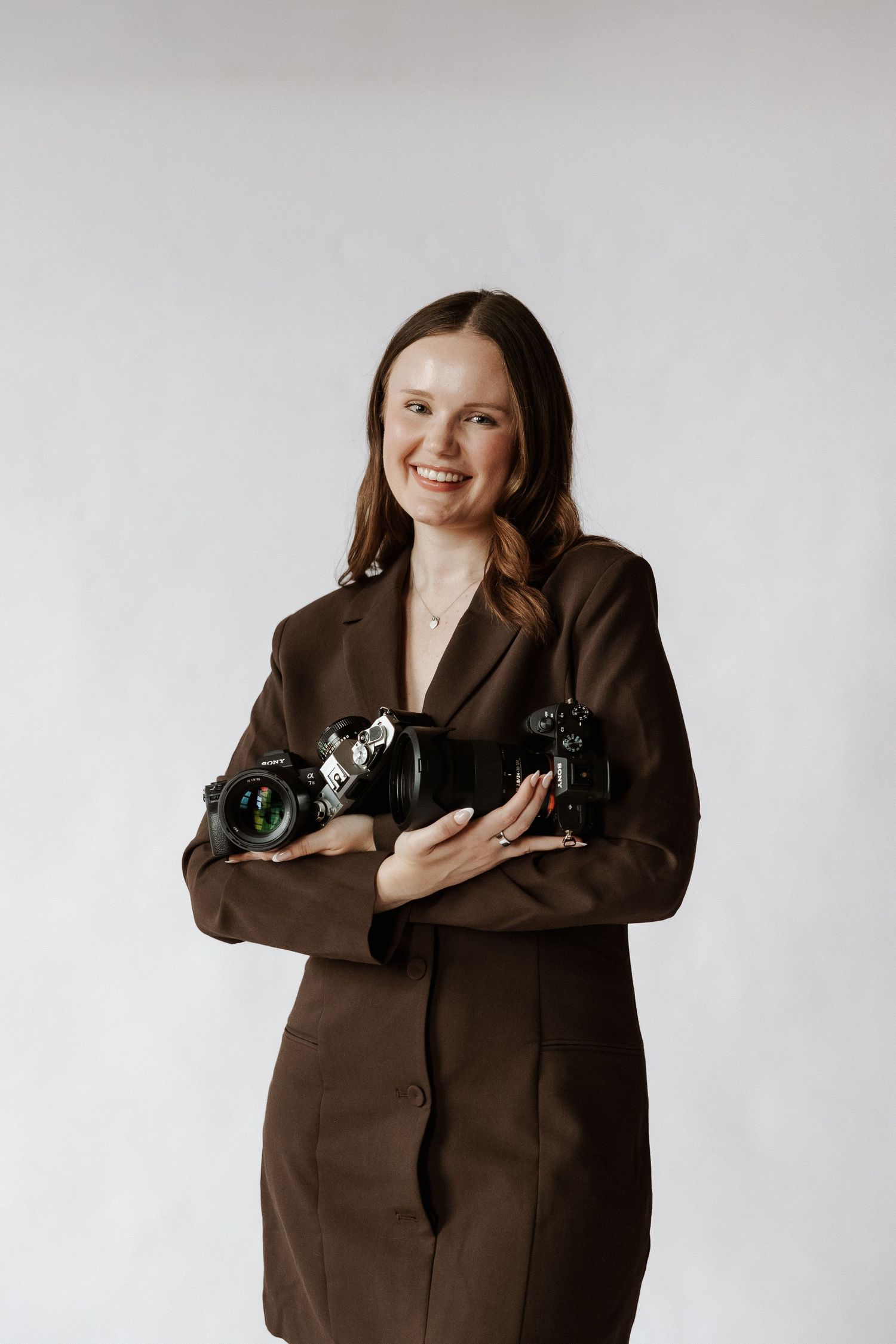 Smiling female photographer in brown blazer holding two professional cameras against white background.