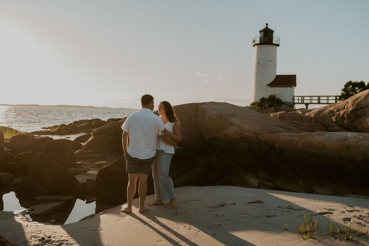 A couple stands on rocky coast at sunset near Annisquam Lighthouse in Gloucester, Massachusetts.