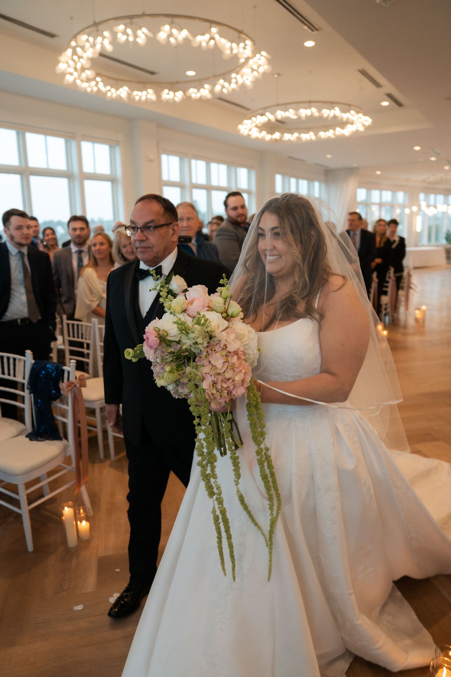 Bride walking down aisle with escort holding pink bouquet in elegant white wedding venue.