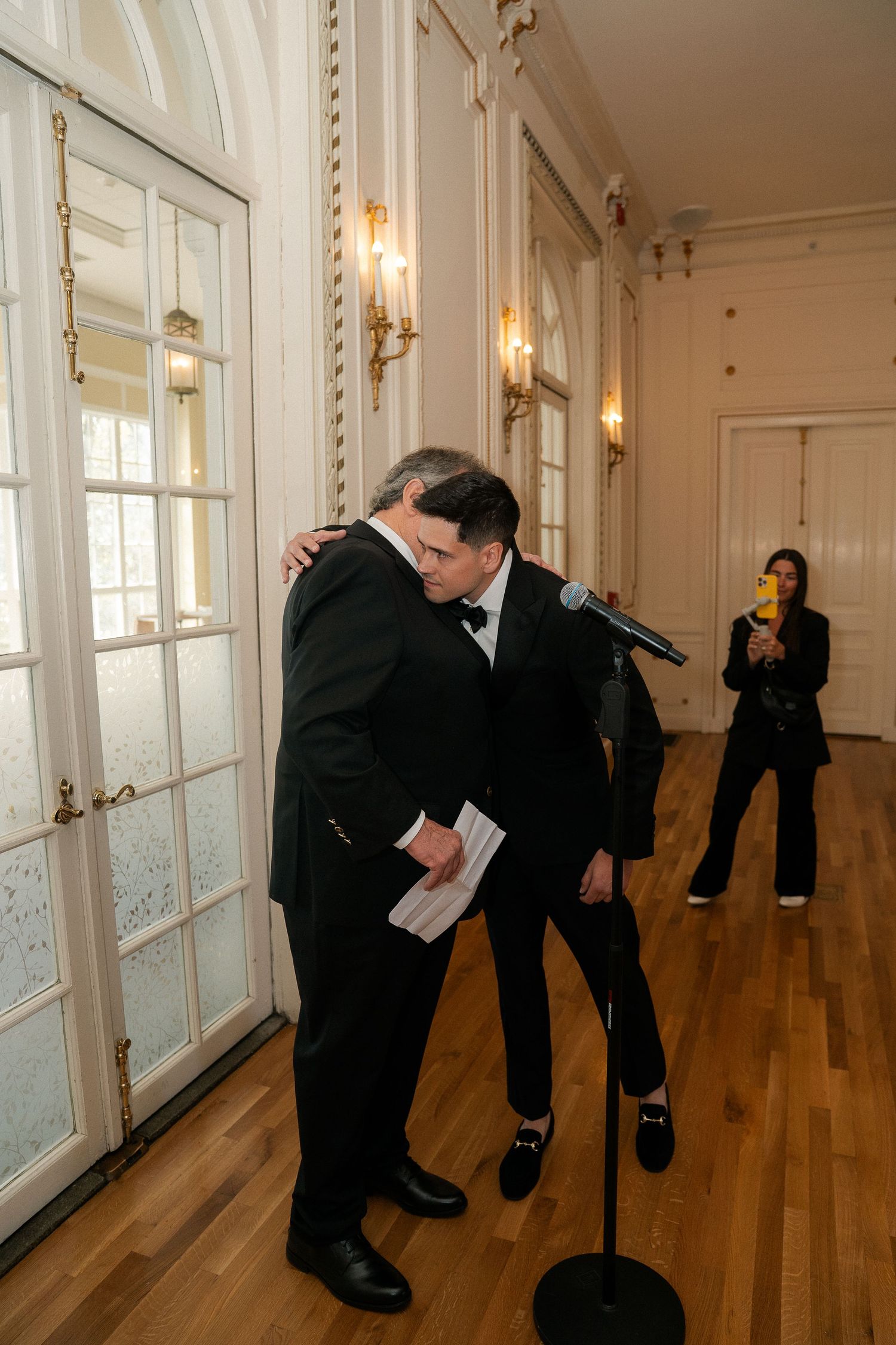 Two men in tuxedos share an emotional hug at a microphone stand in an elegant white ballroom.
