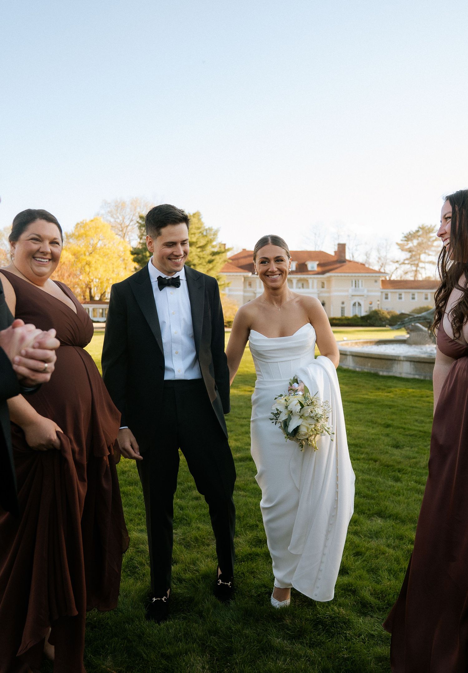 Bride and groom smiling outdoors with bridesmaids in brown dresses at elegant estate wedding venue.