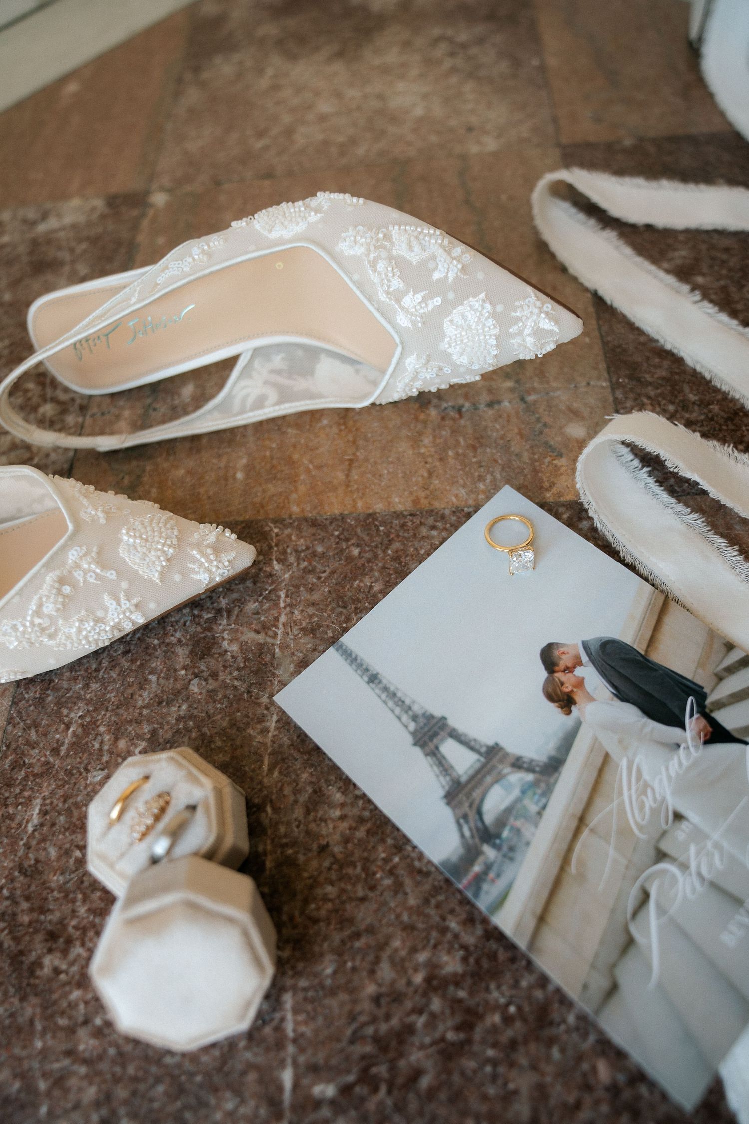 Bridal flat shoes with lace detail, gold rings in velvet box, and Eiffel Tower photo on marble floor.