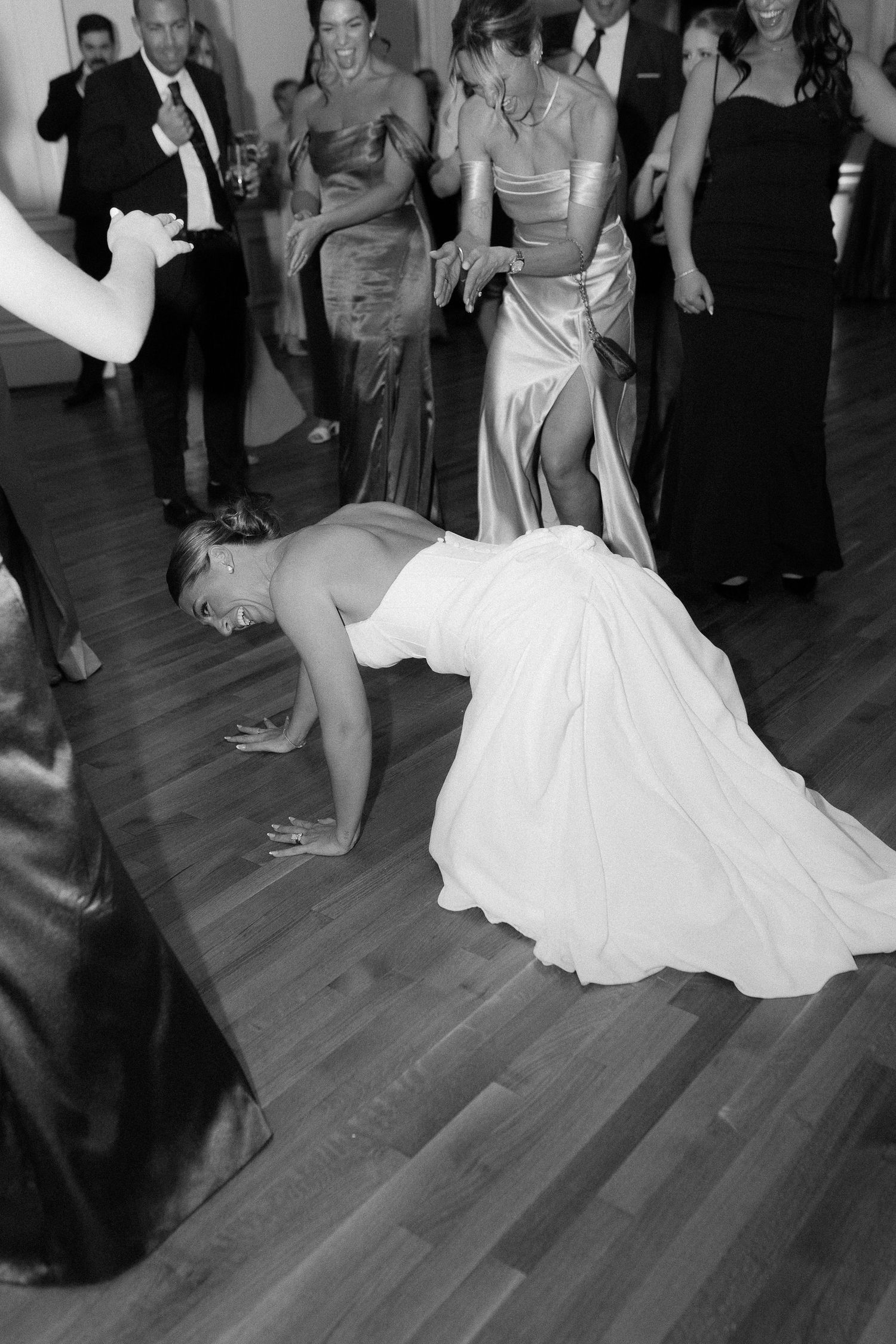 Bride in white wedding gown doing push-ups on dance floor surrounded by guests at lively reception.