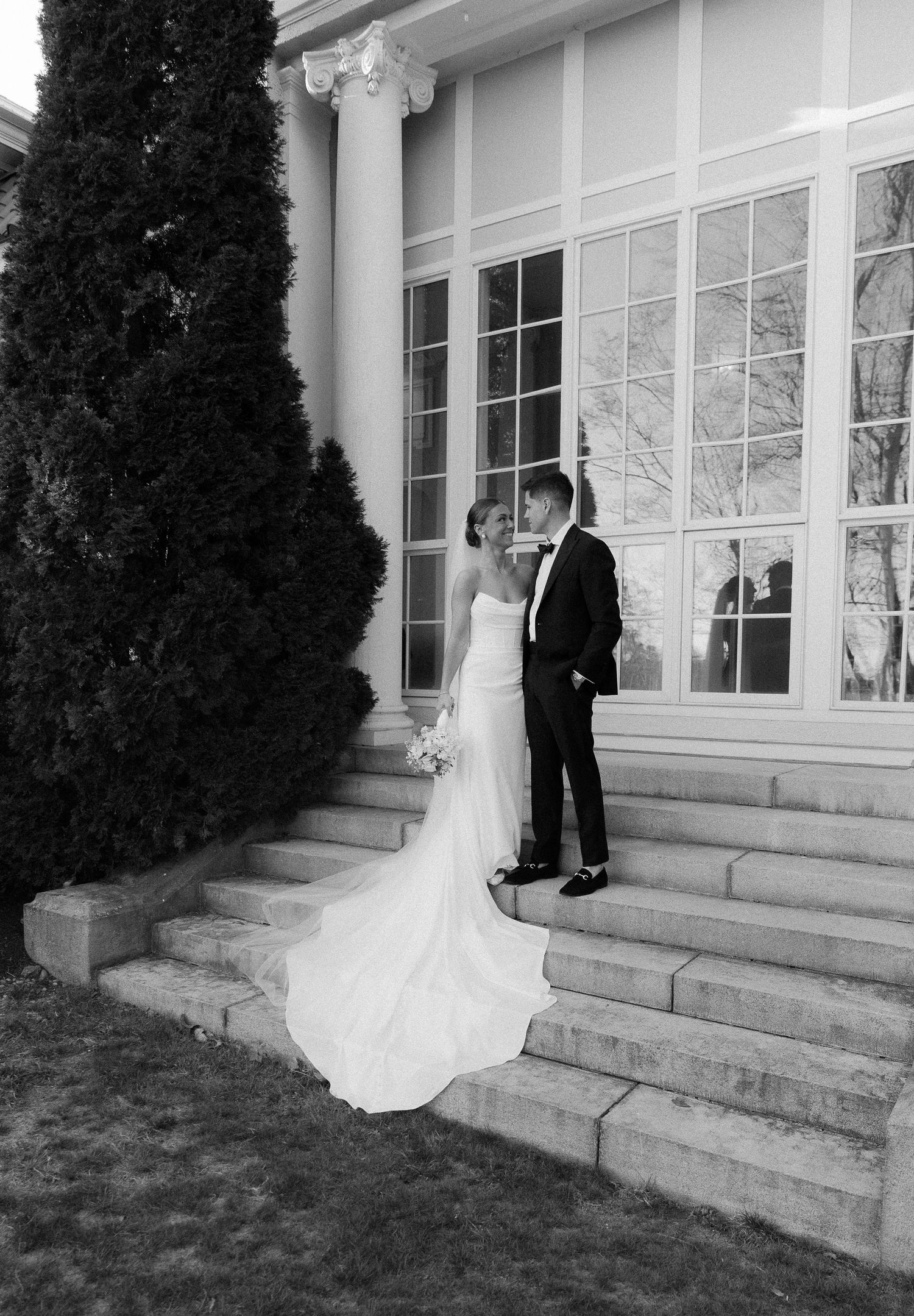 Bride and groom share intimate moment on stone steps beside tall cypress trees.