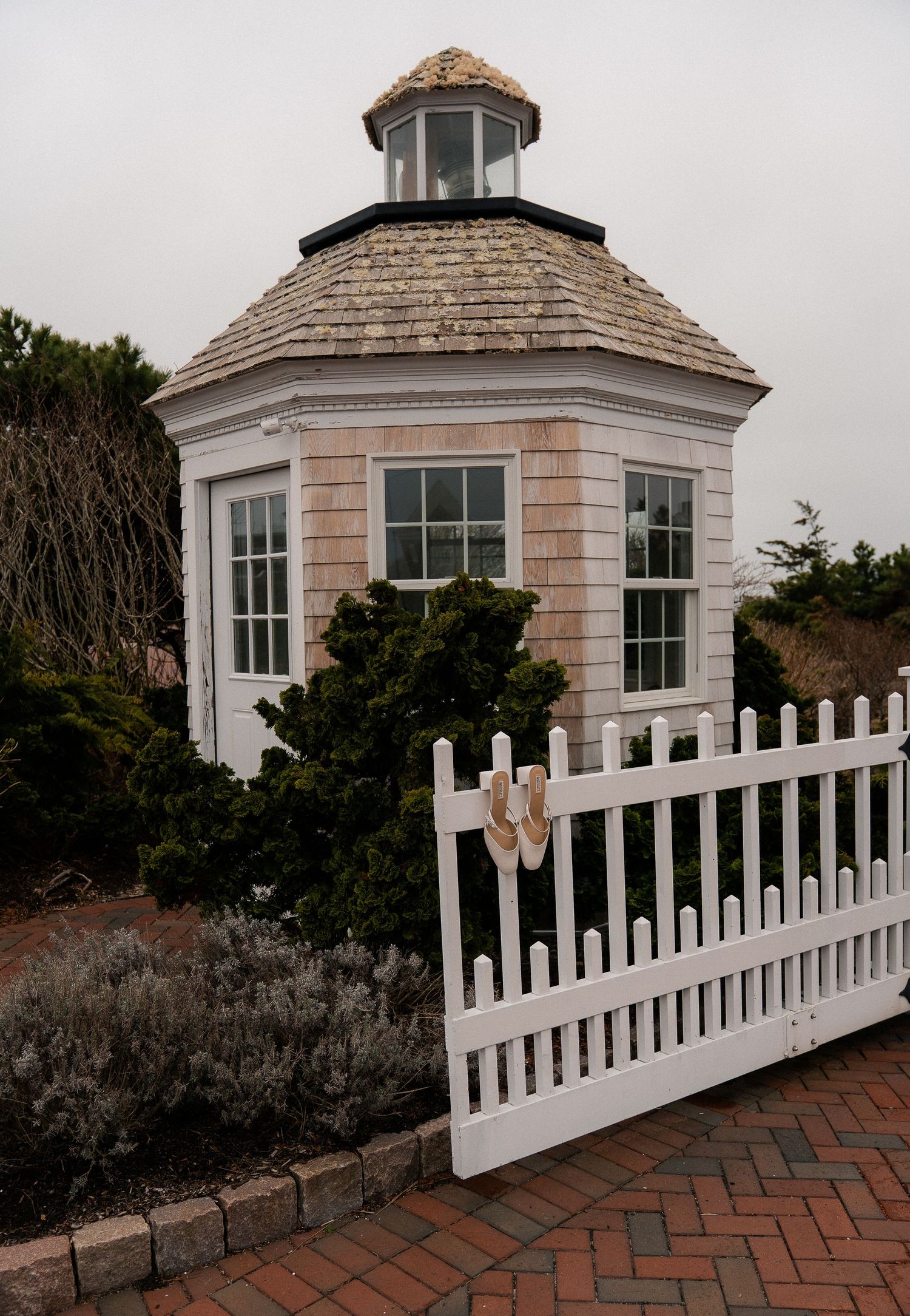 Charming octagonal gazebo with cedar shake roof and white picket fence gate on a brick patio.