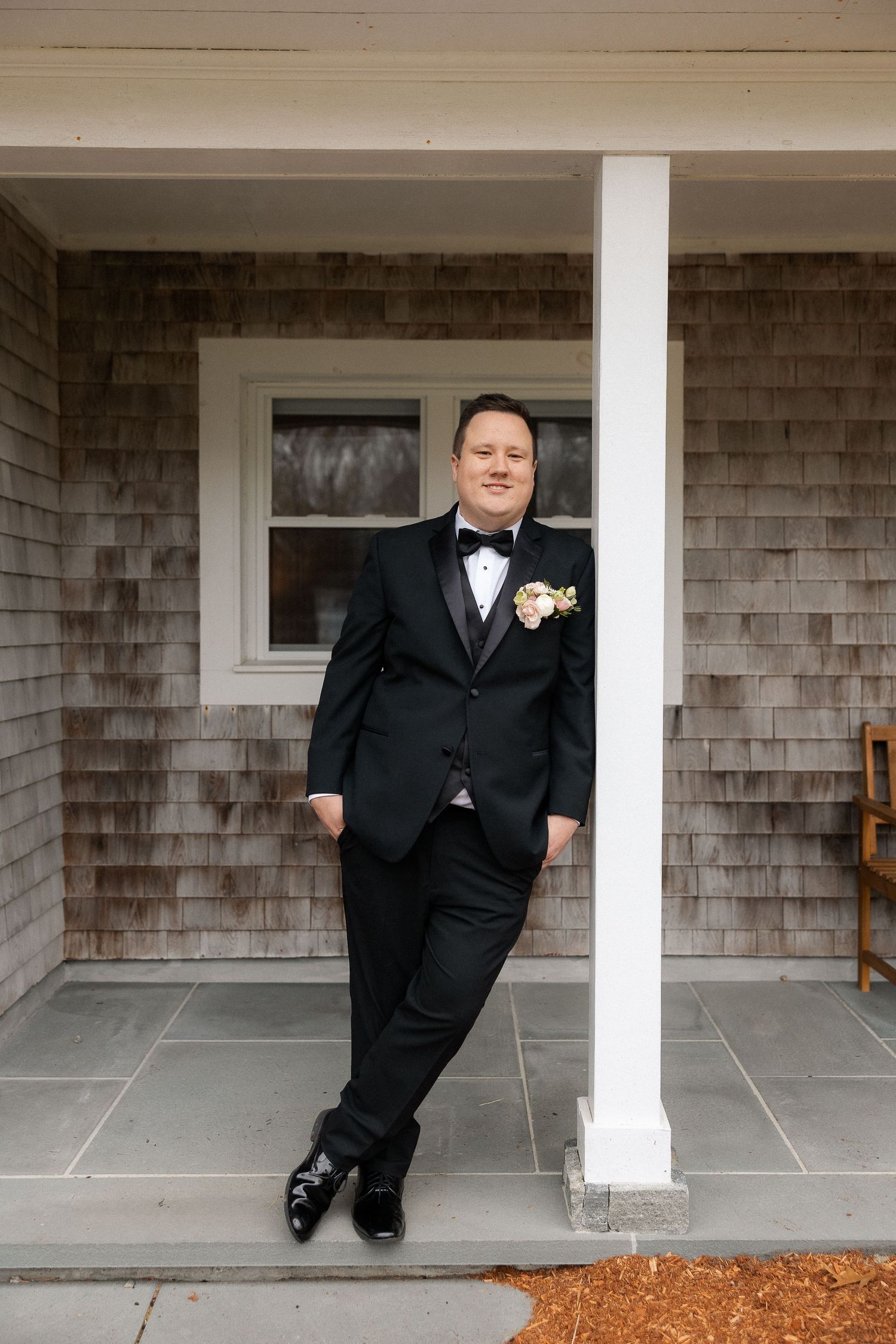 Dapper groom in a black tuxedo with floral boutonniere leaning against a white porch column of a shingle-style home.