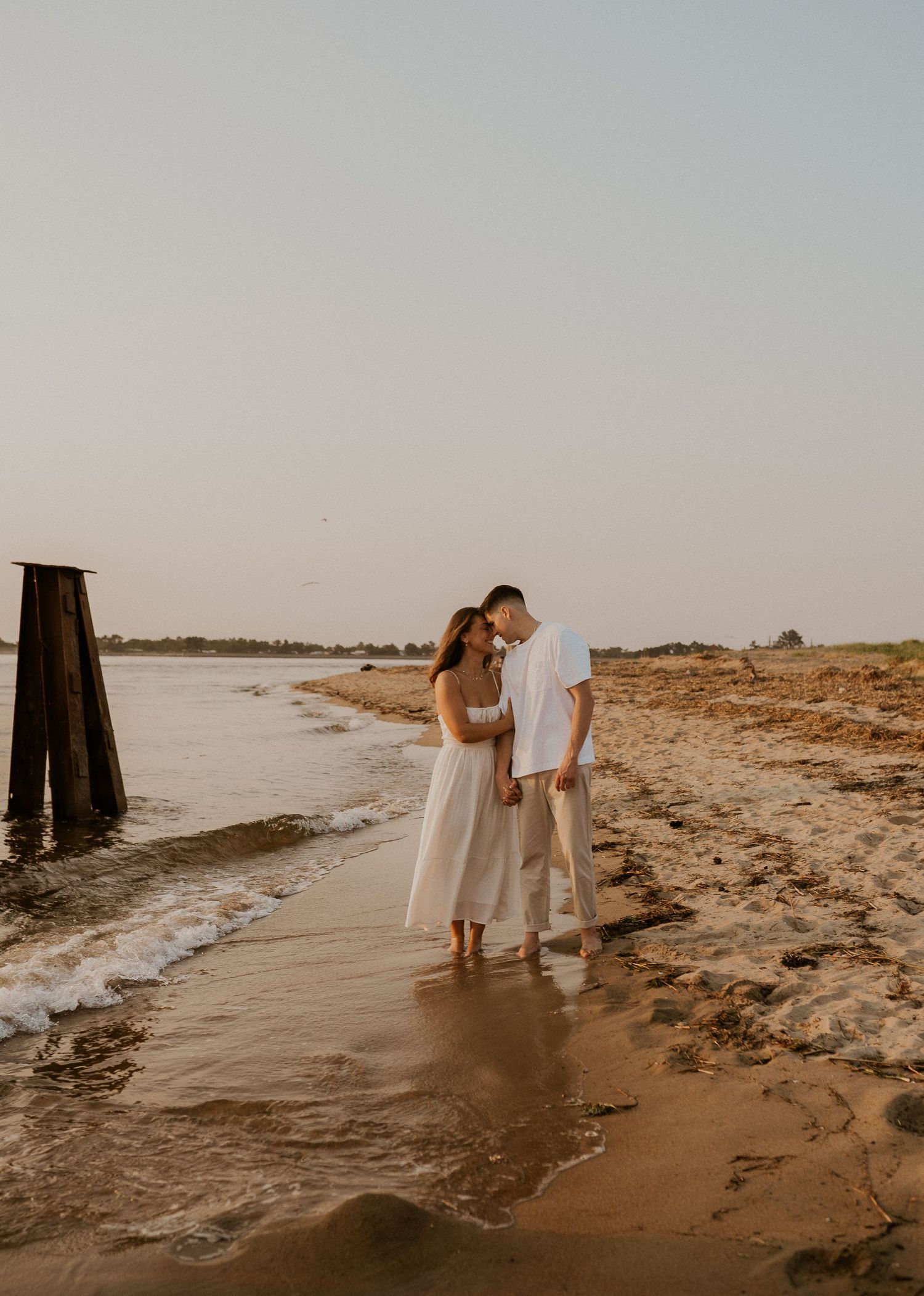 Couple in white outfits walking along the water's edge at a secluded beach during sunset.