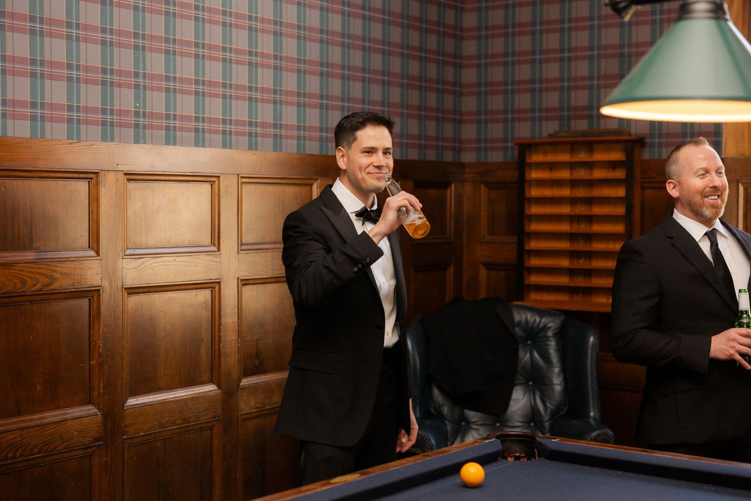 Two men in black tuxedos enjoy drinks in an elegant billiard room with plaid wallpaper and pool table.
