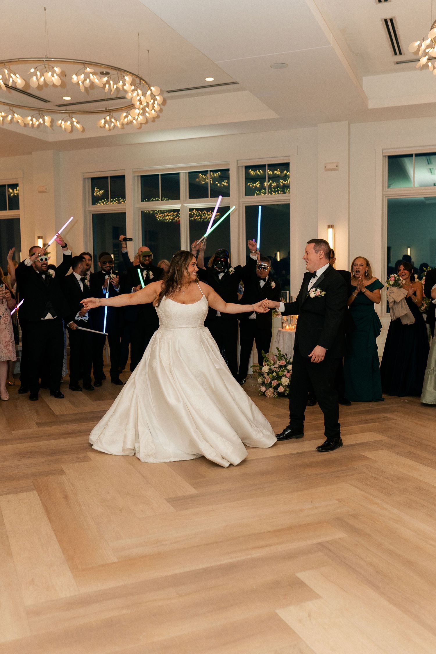Bride in strapless ball gown and groom share first dance surrounded by guests holding light sabers at wedding reception.