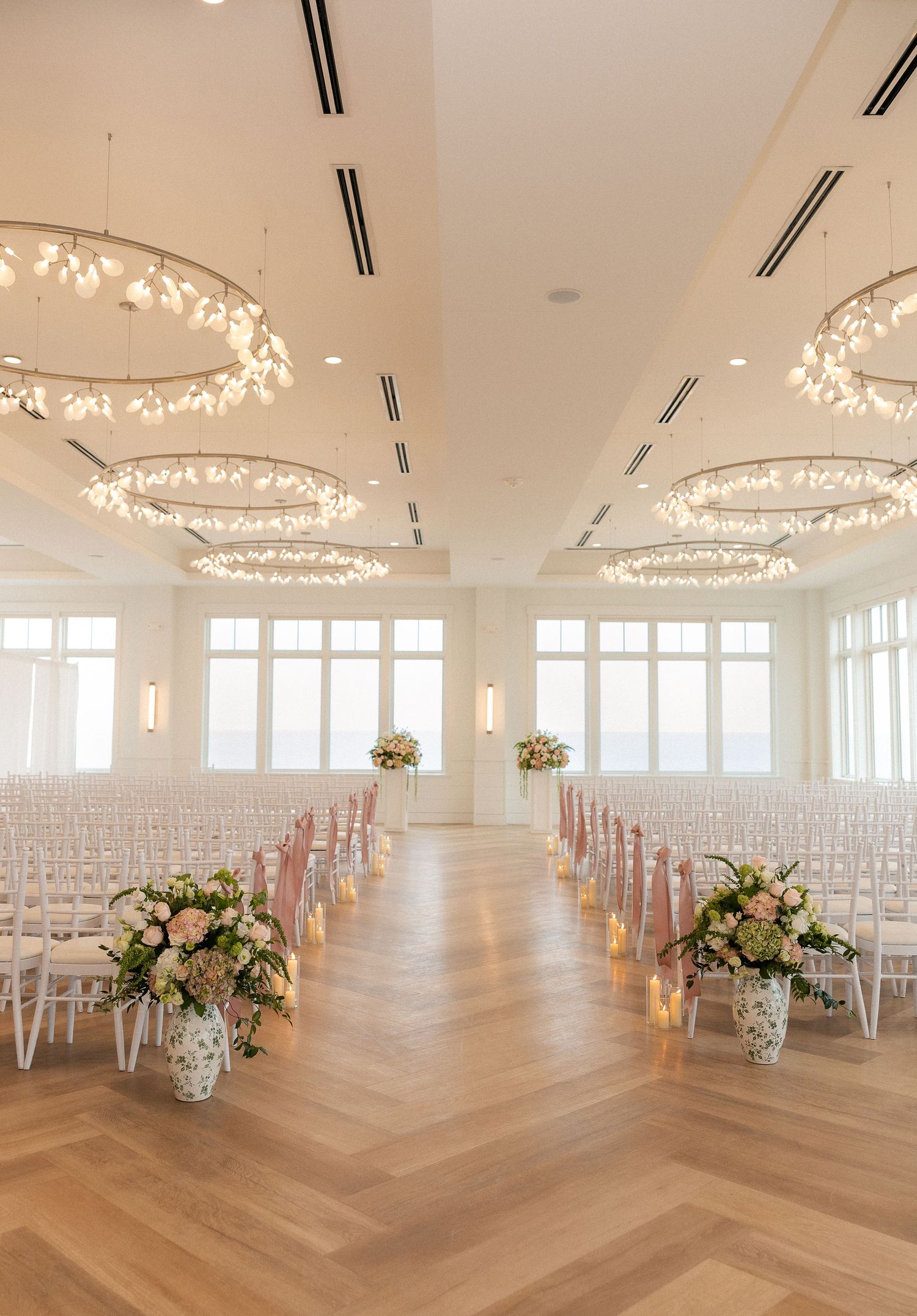 Elegant wedding ceremony venue with white chairs, floral arrangements, herringbone floors, and circular chandeliers in a bright ballroom.