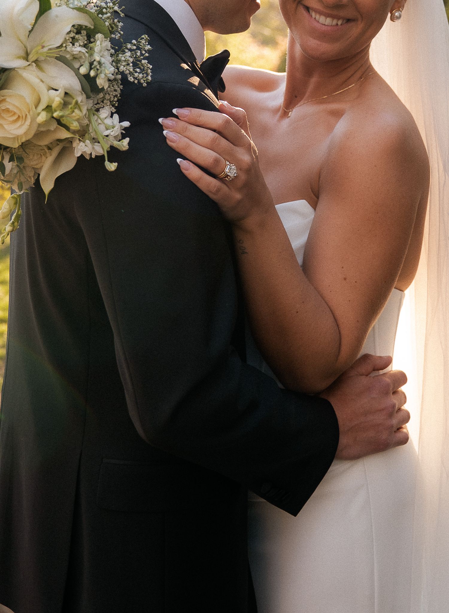 Bride and groom embracing outdoors, bride wearing strapless white gown and diamond ring, holding white floral bouquet.