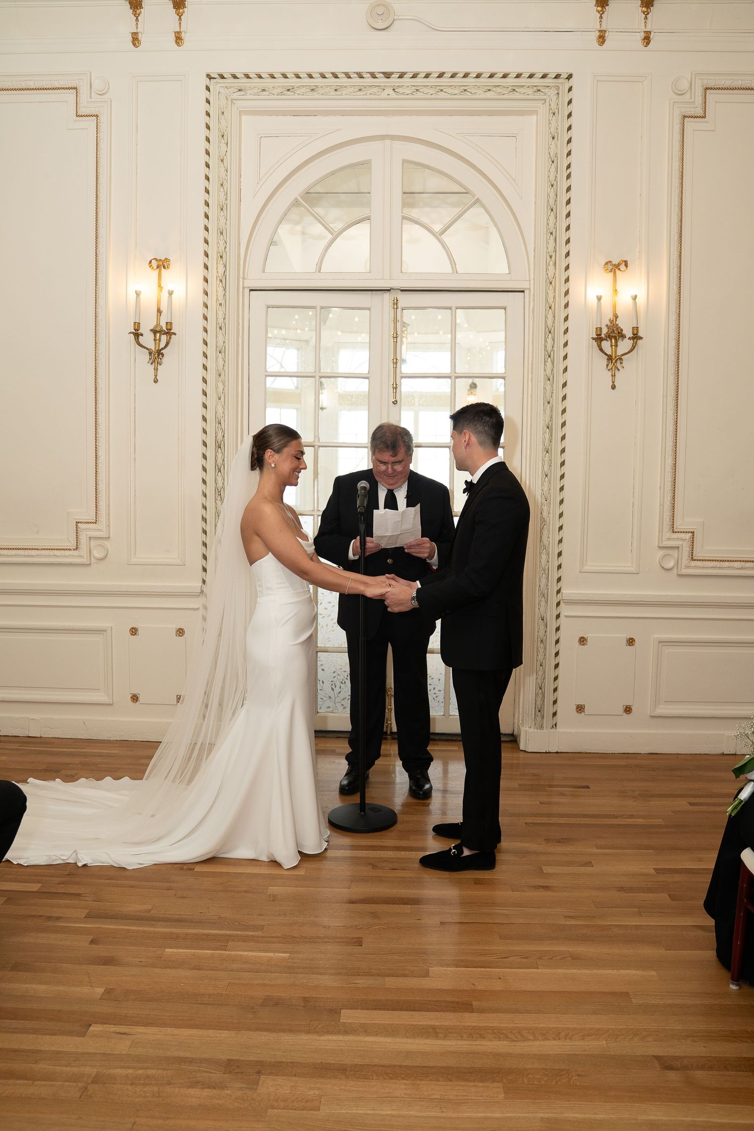 Bride and groom exchange vows with officiant in elegant white ballroom during indoor wedding ceremony.