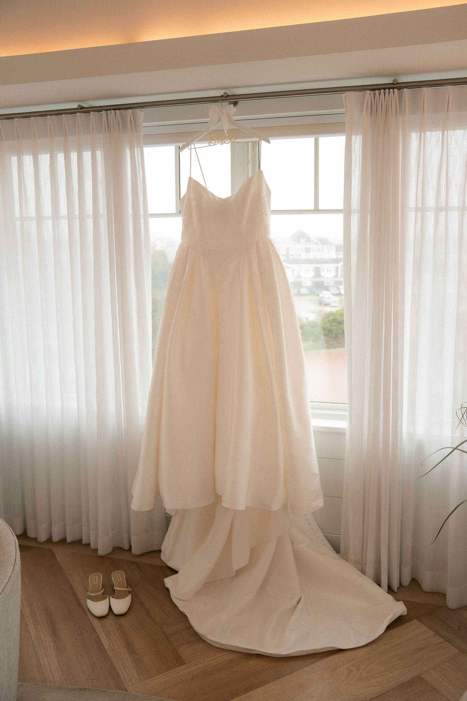 Elegant ivory wedding gown with a long train hanging by a sunlit window with sheer curtains and bridal heels below.